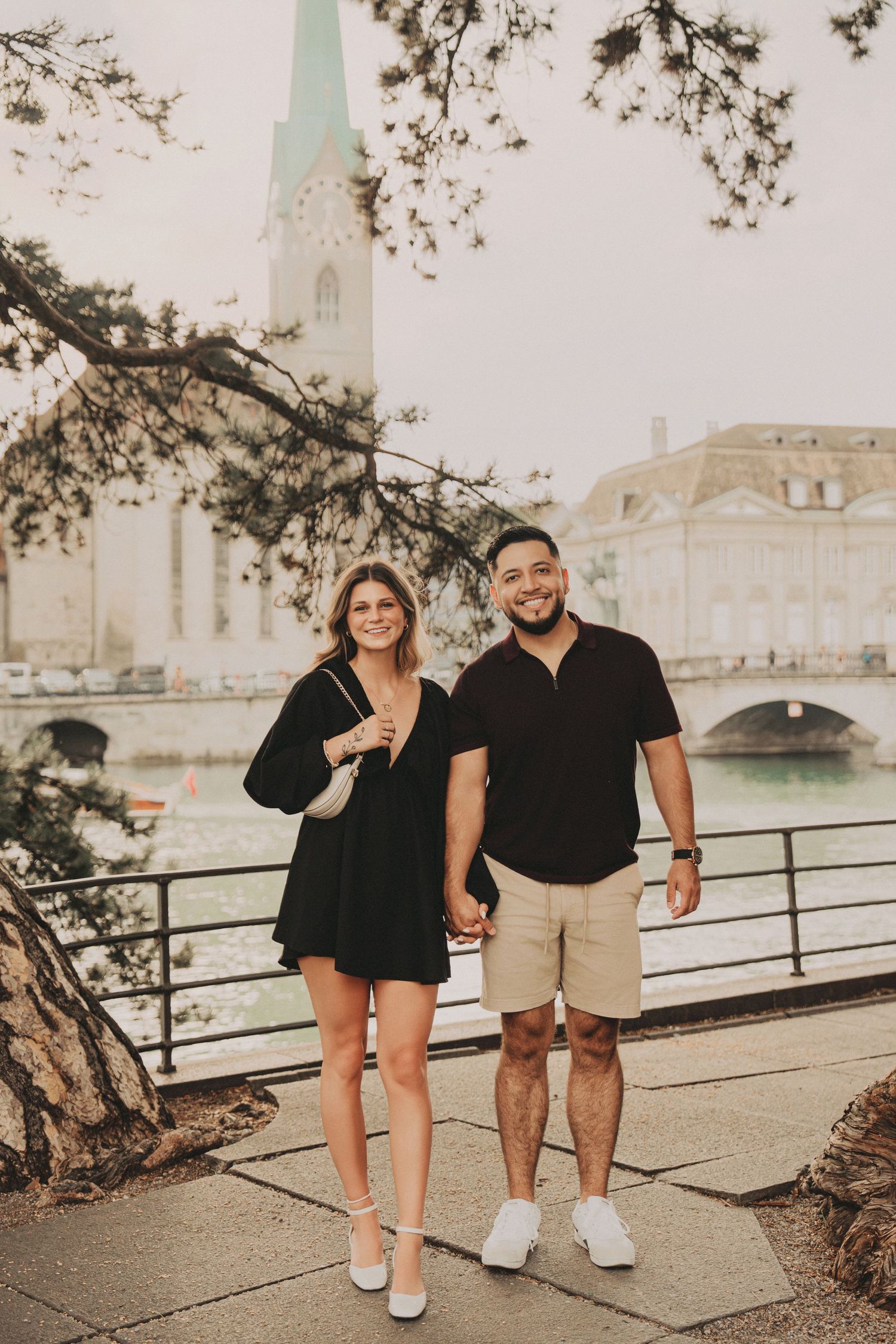 Smiling couple holding hands in black outfits by Zurich river with Fraumunster church backdrop.