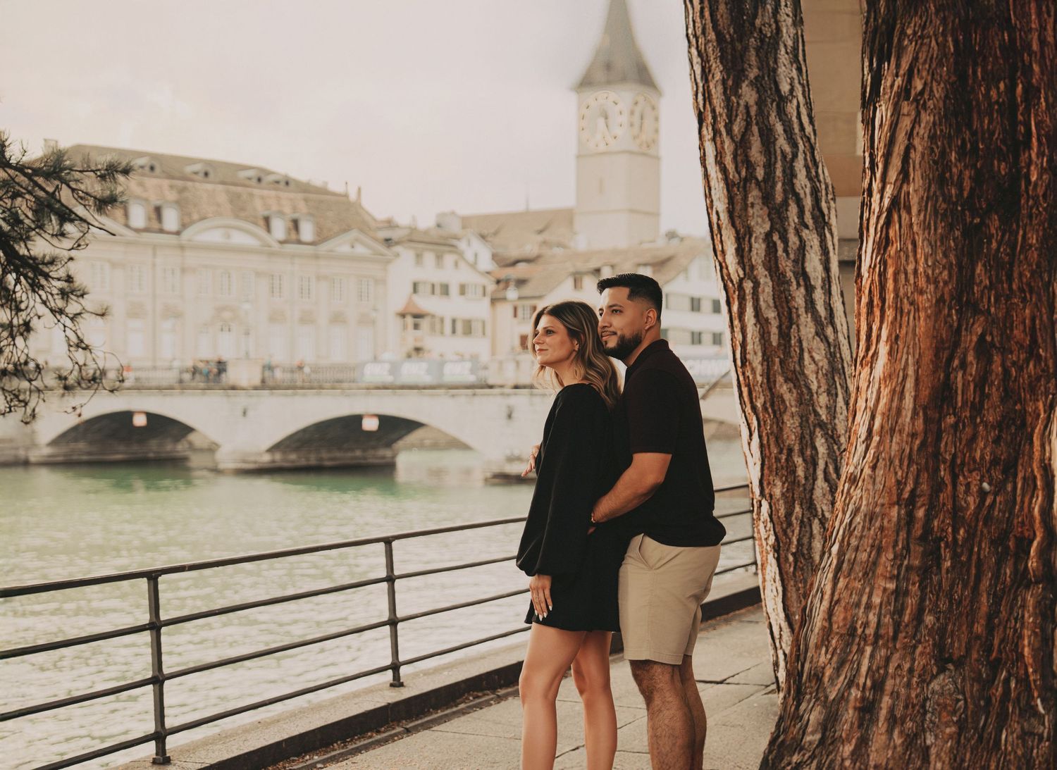 Couple in black outfits standing by Zurich riverfront with historic bridge and tower in background.