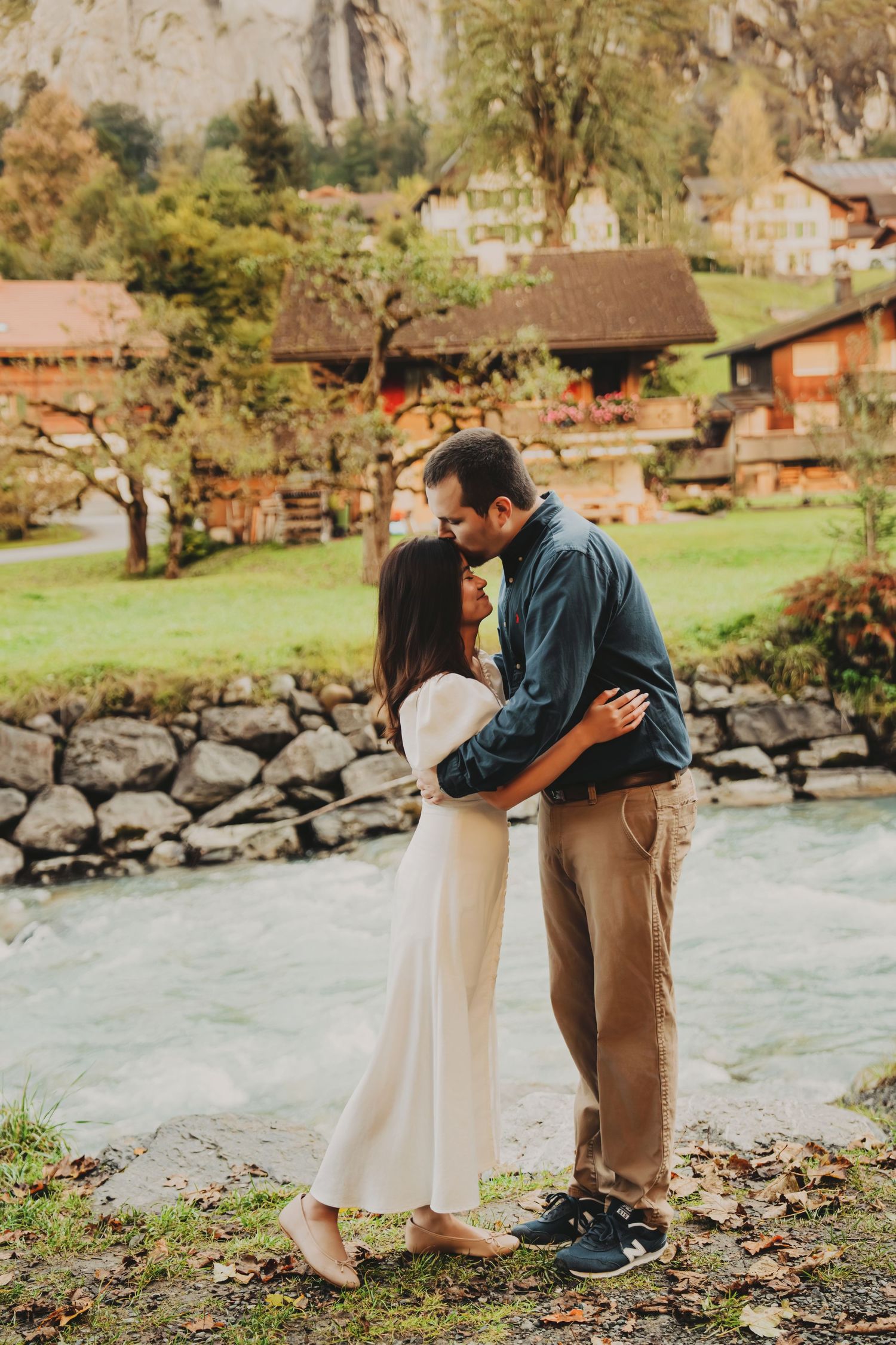 Romantic couple kissing near a stream with charming Swiss chalet village backdrop.