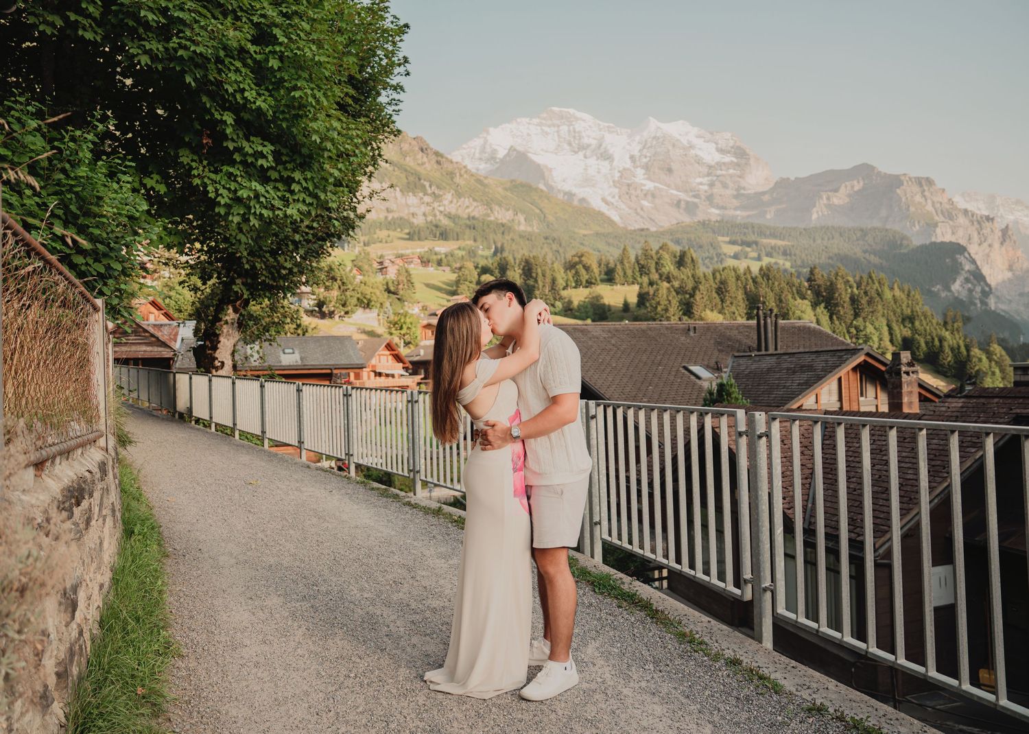 Couple kissing on a picturesque Swiss village path with dramatic snow-covered mountain peaks in the background.