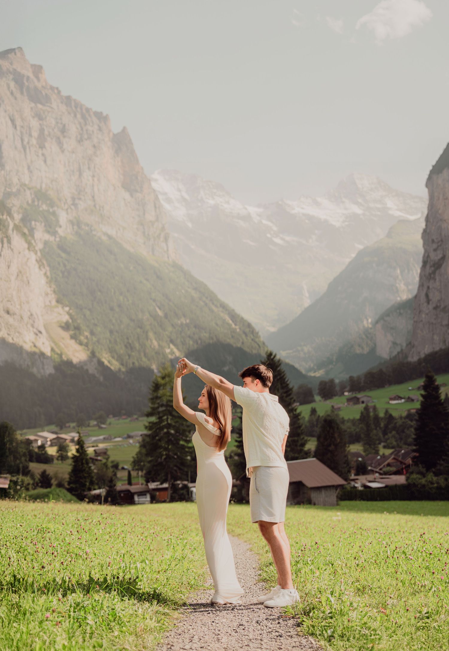 Couple dancing joyfully on a grassy alpine path with spectacular snow-capped Swiss mountain peaks in the background.