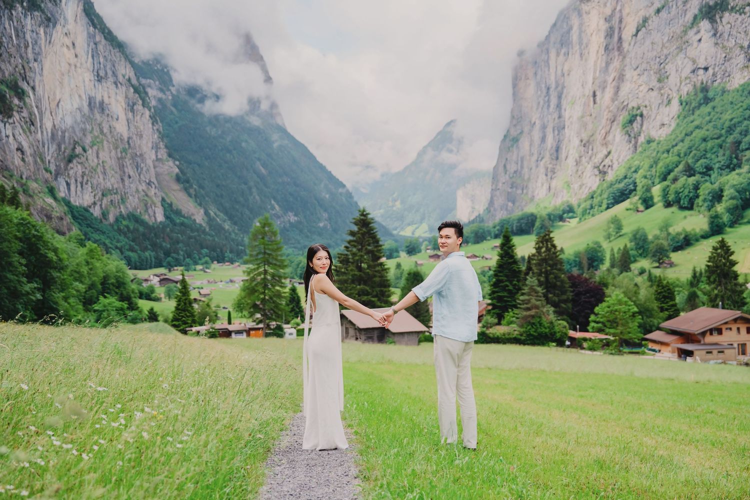 Couple holds hands on a scenic path in Lauterbrunnen valley, Switzerland, with dramatic cliffs and green meadows.