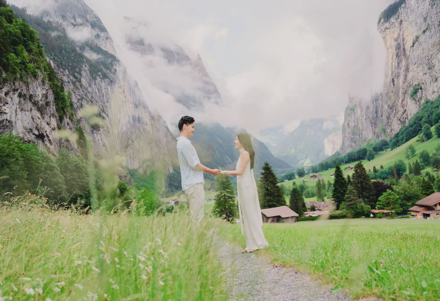 Couple holding hands on a scenic mountain path in Lauterbrunnen valley, Switzerland, surrounded by lush greenery and misty peaks.