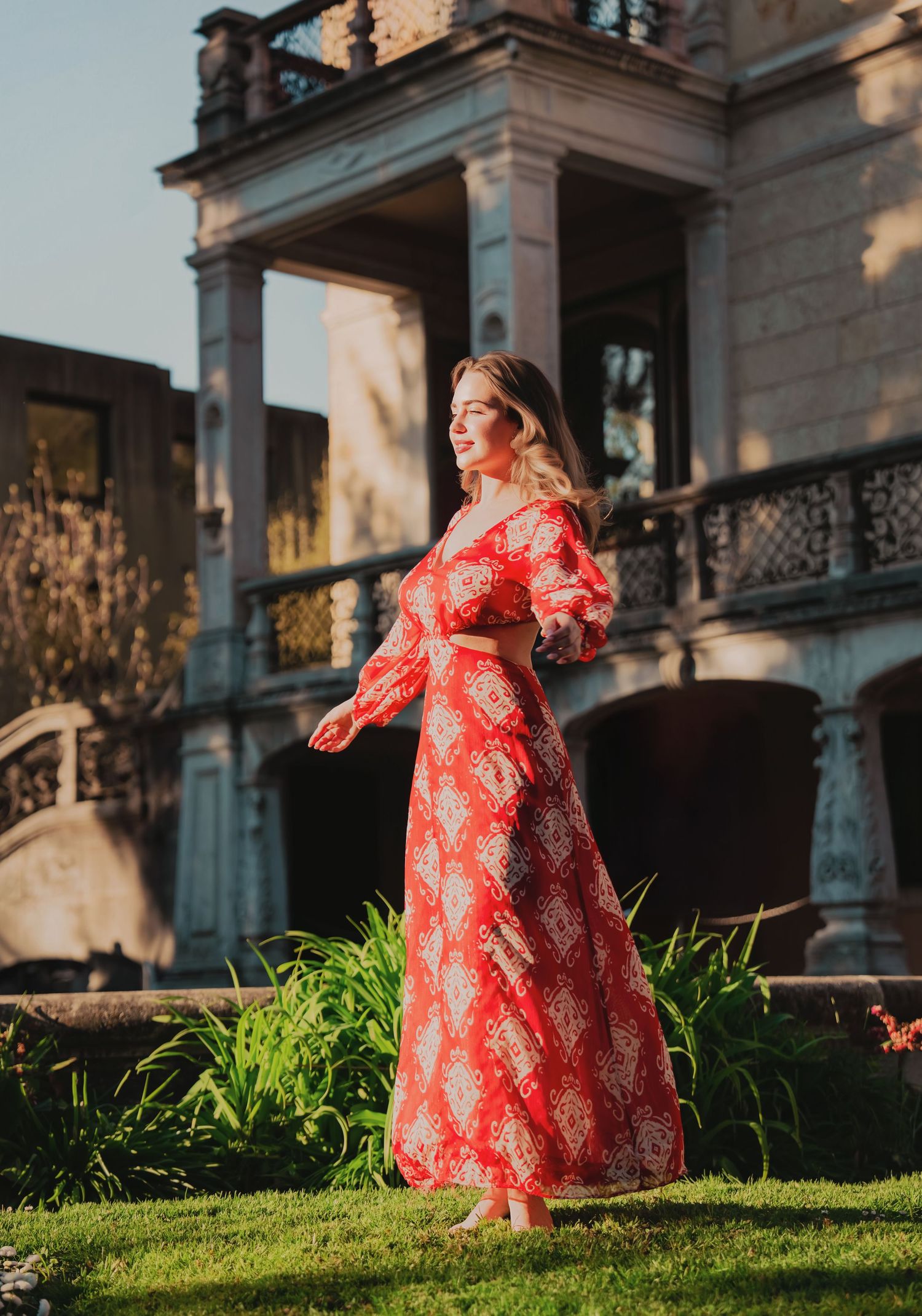 Elegant woman in red patterned maxi dress stands before ornate historic mansion in golden sunlight.