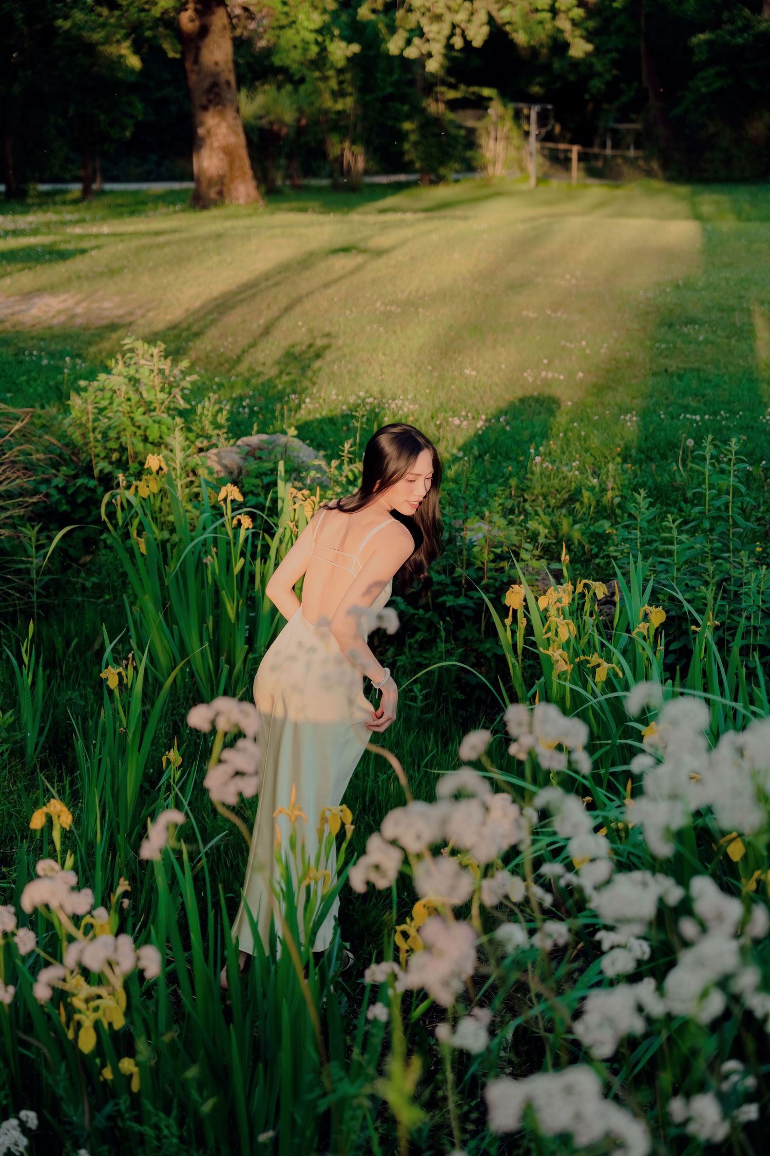 Woman in sheer dress standing among wildflowers and iris blooms in a sunlit green garden at golden hour.