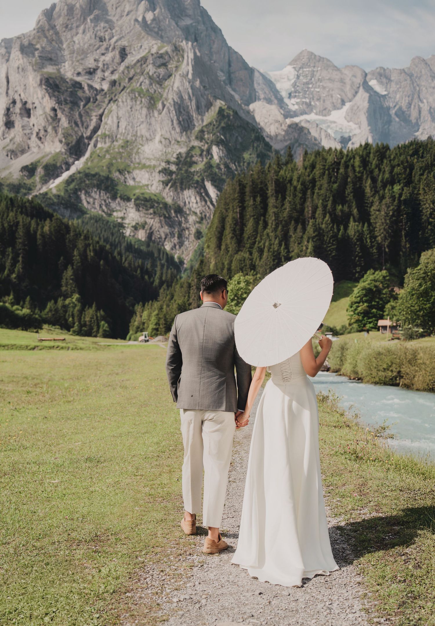 Wedding couple strolling riverside path with bride holding white parasol and mountain scenery.