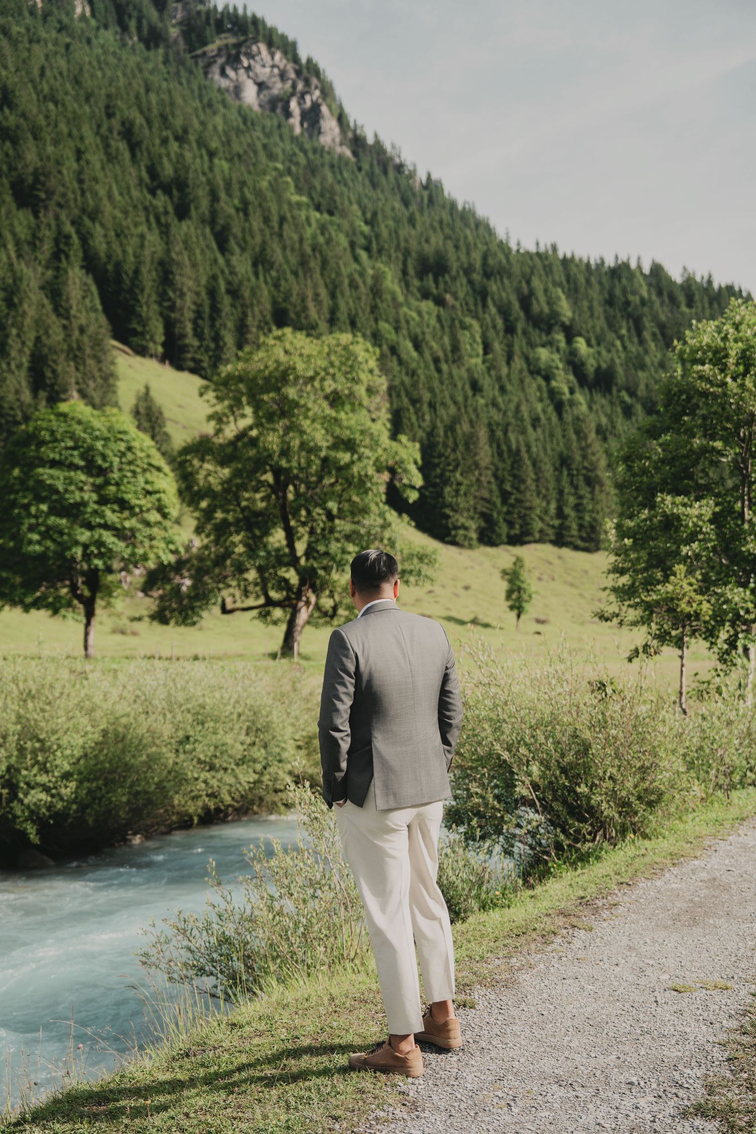 Groom in grey blazer and cream trousers walks along mountain stream path surrounded by lush green forest.