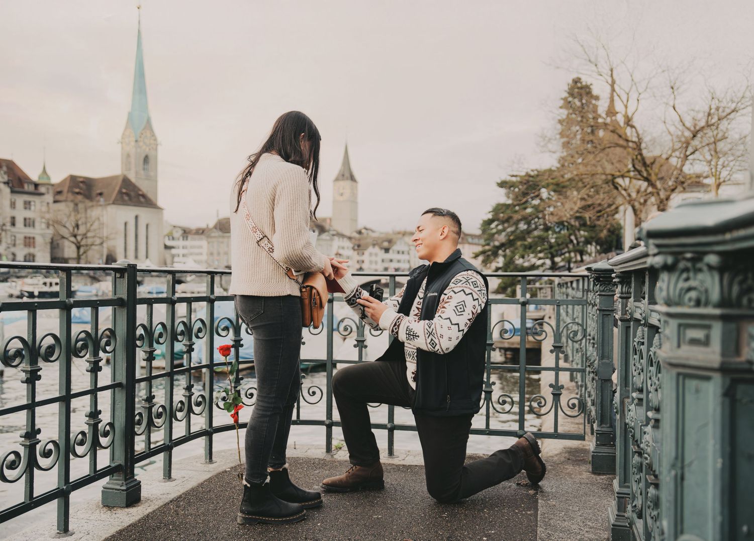 Man down on one knee proposing to woman on scenic Zurich bridge with church towers.