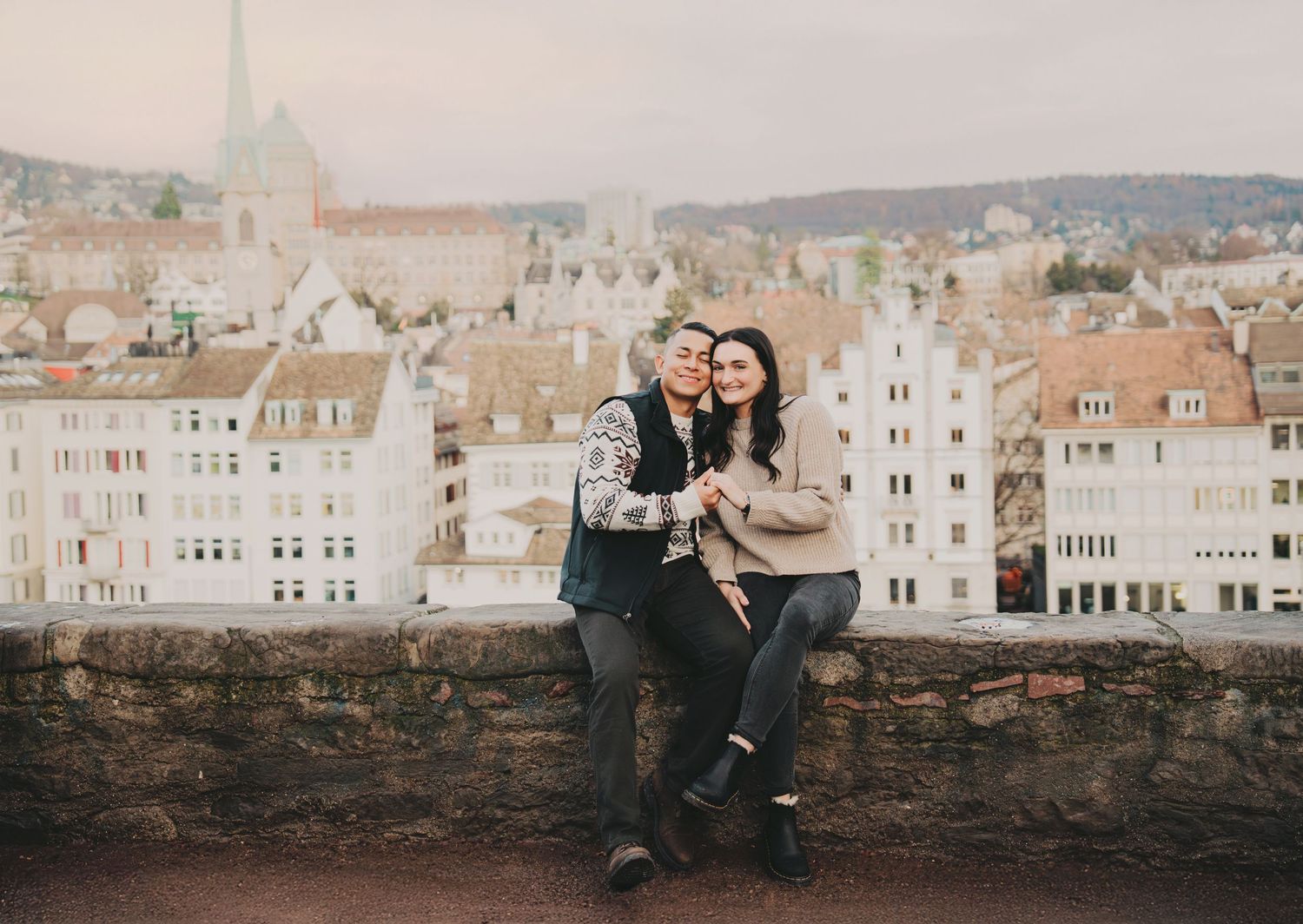 Couple cuddling and laughing on a stone wall with a European city skyline behind them.