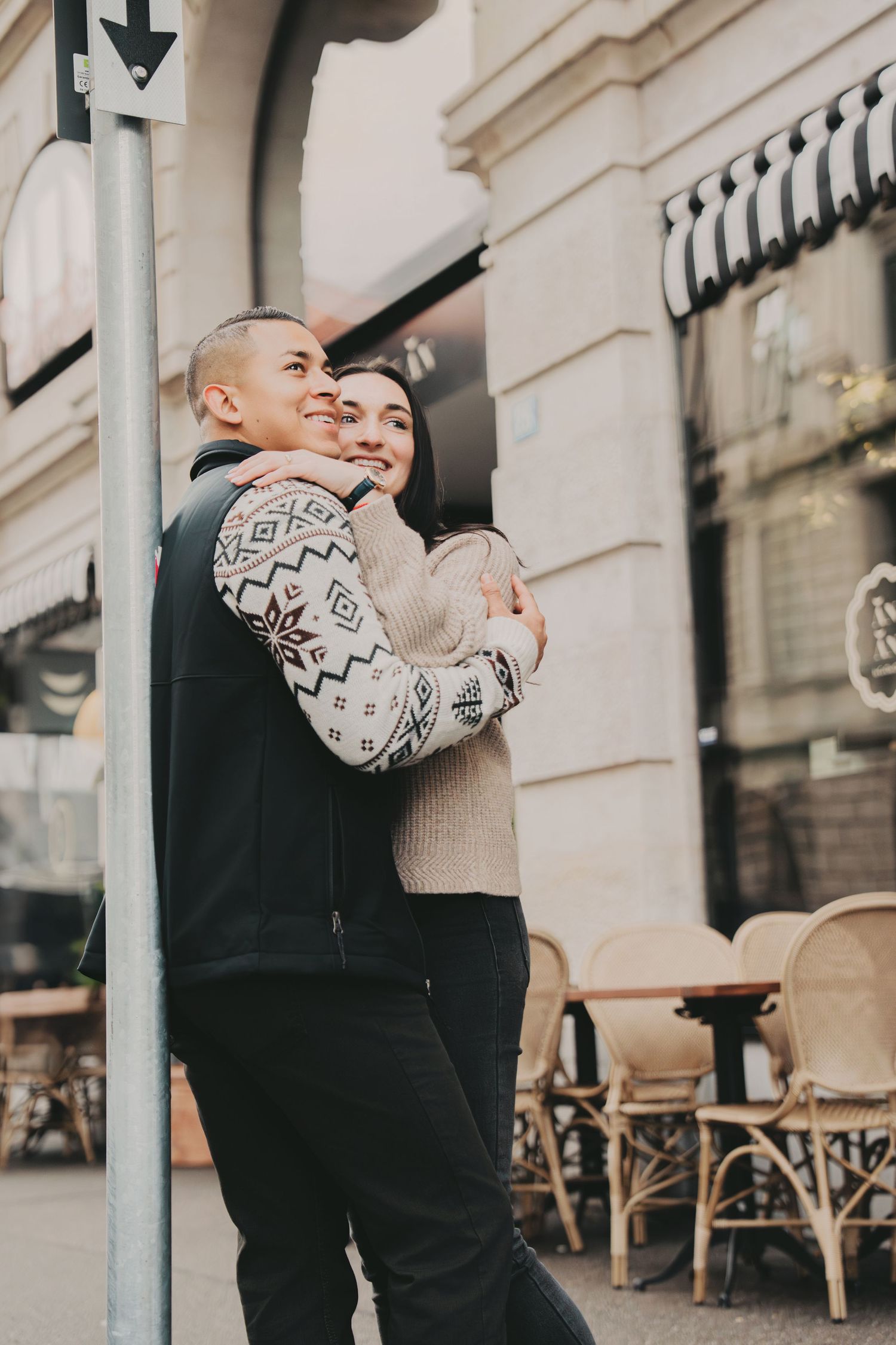 Happy couple embracing and smiling outside a European cafe with bistro chairs nearby.