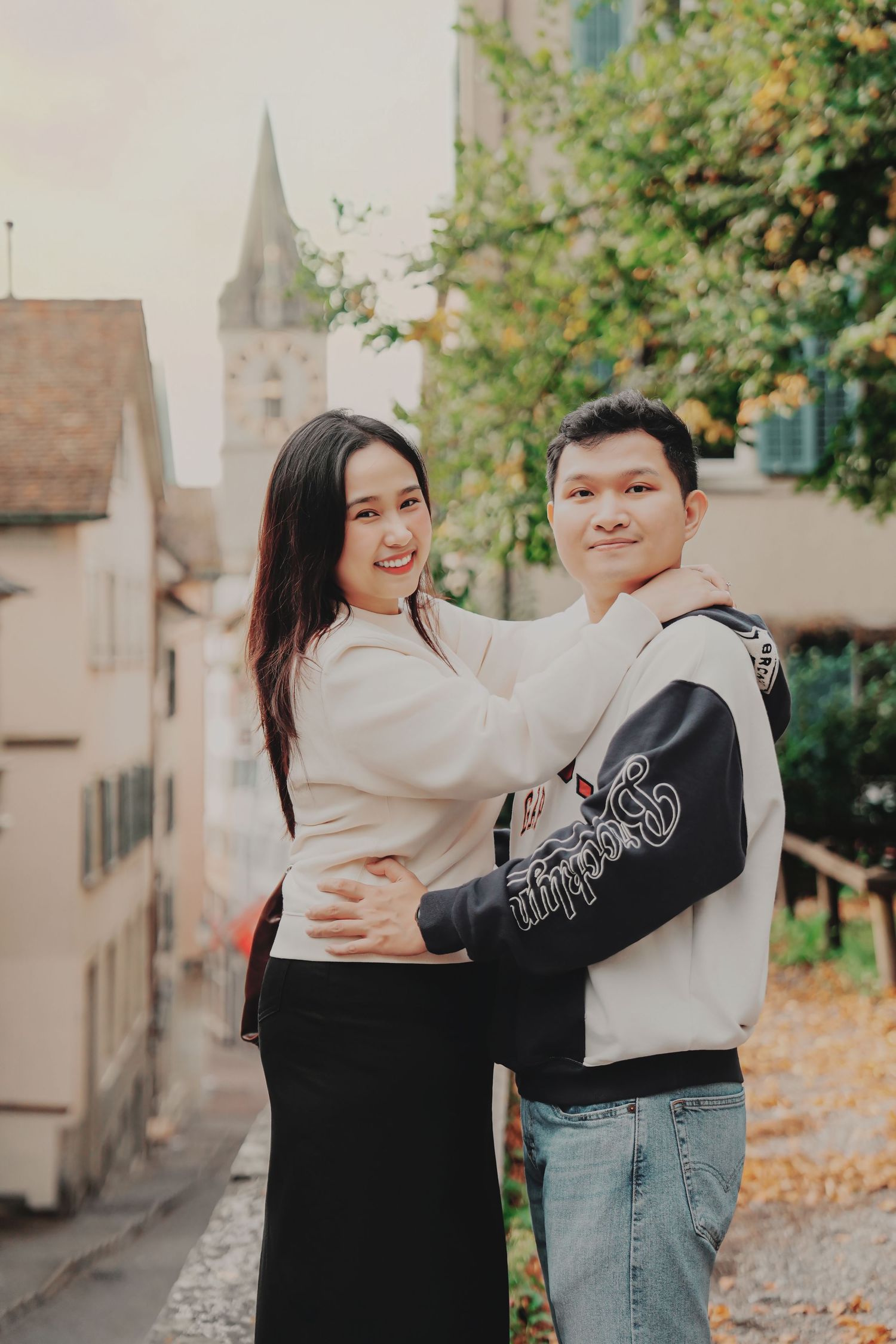 Smiling Asian couple embracing on a tree-lined street in Zurich old town in autumn.