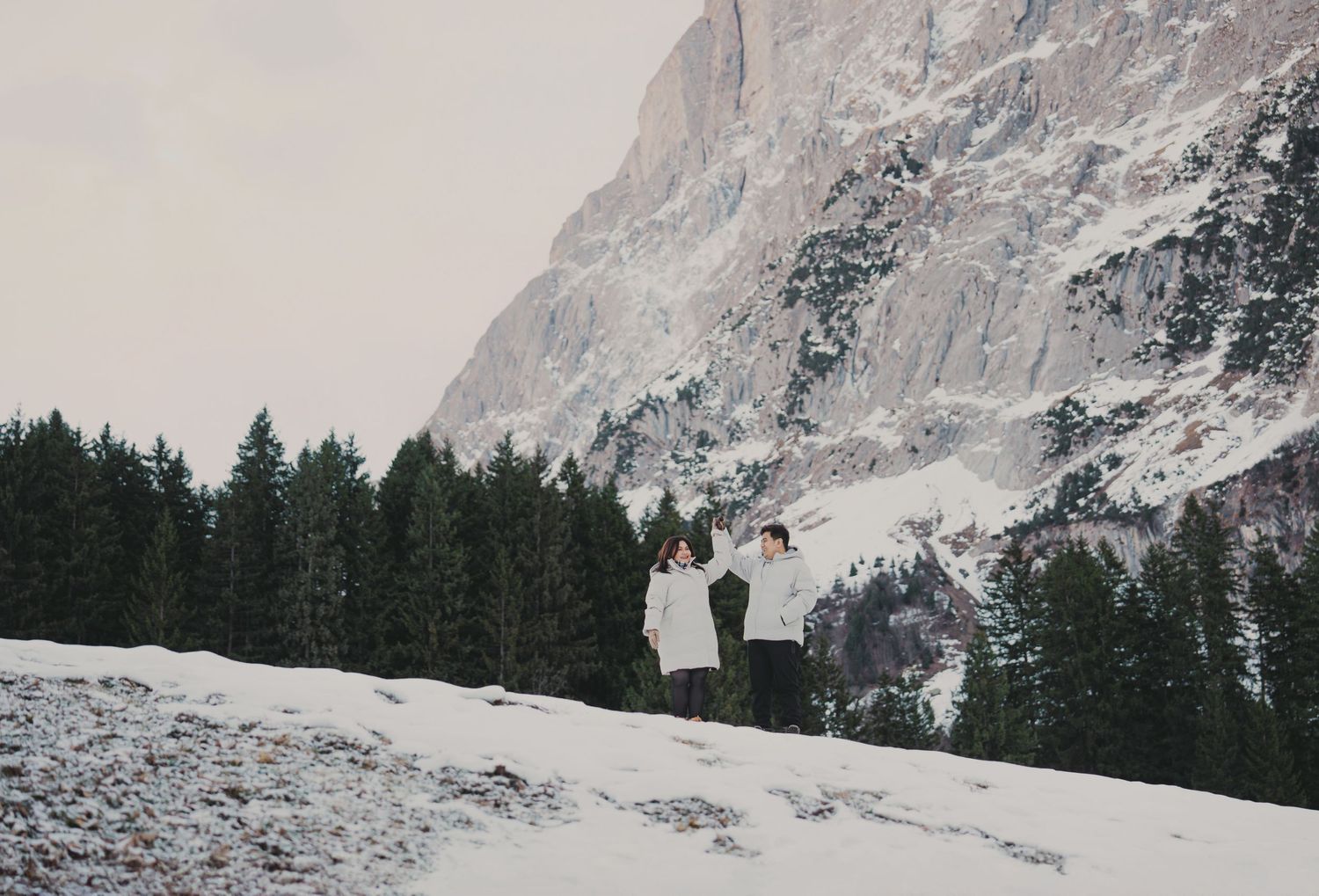 Couple posing playfully in snow with tall evergreen trees and snowy mountain behind them.