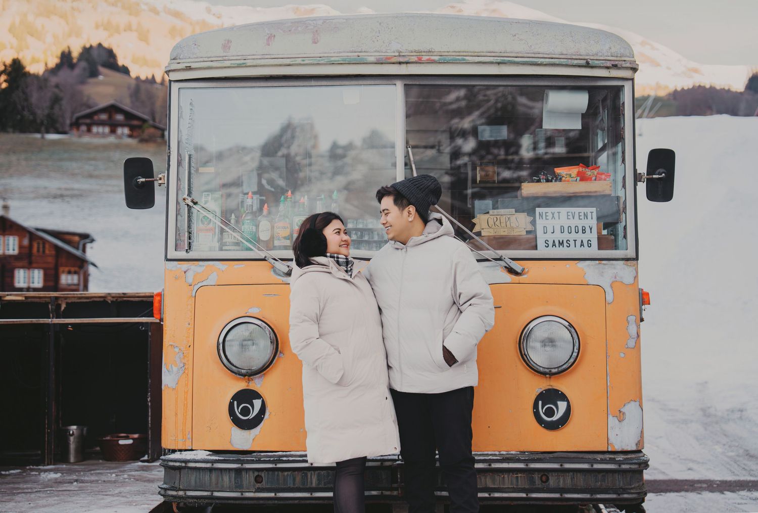 Couple standing together in front of vintage yellow bus in snowy alpine setting.