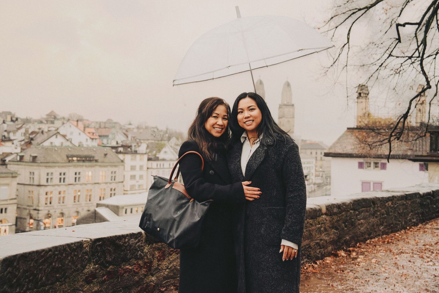 Mom and daughter smiling together under a clear umbrella with the Zurich skyline on a rainy day.