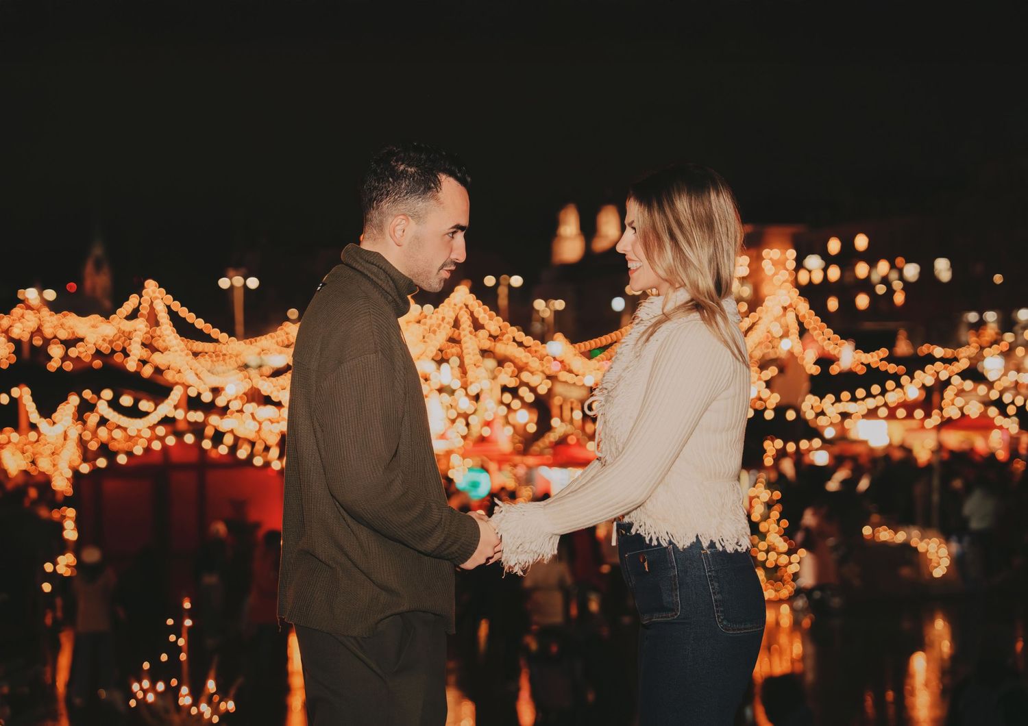 Couple holding hands at night surrounded by glowing Christmas market lights.