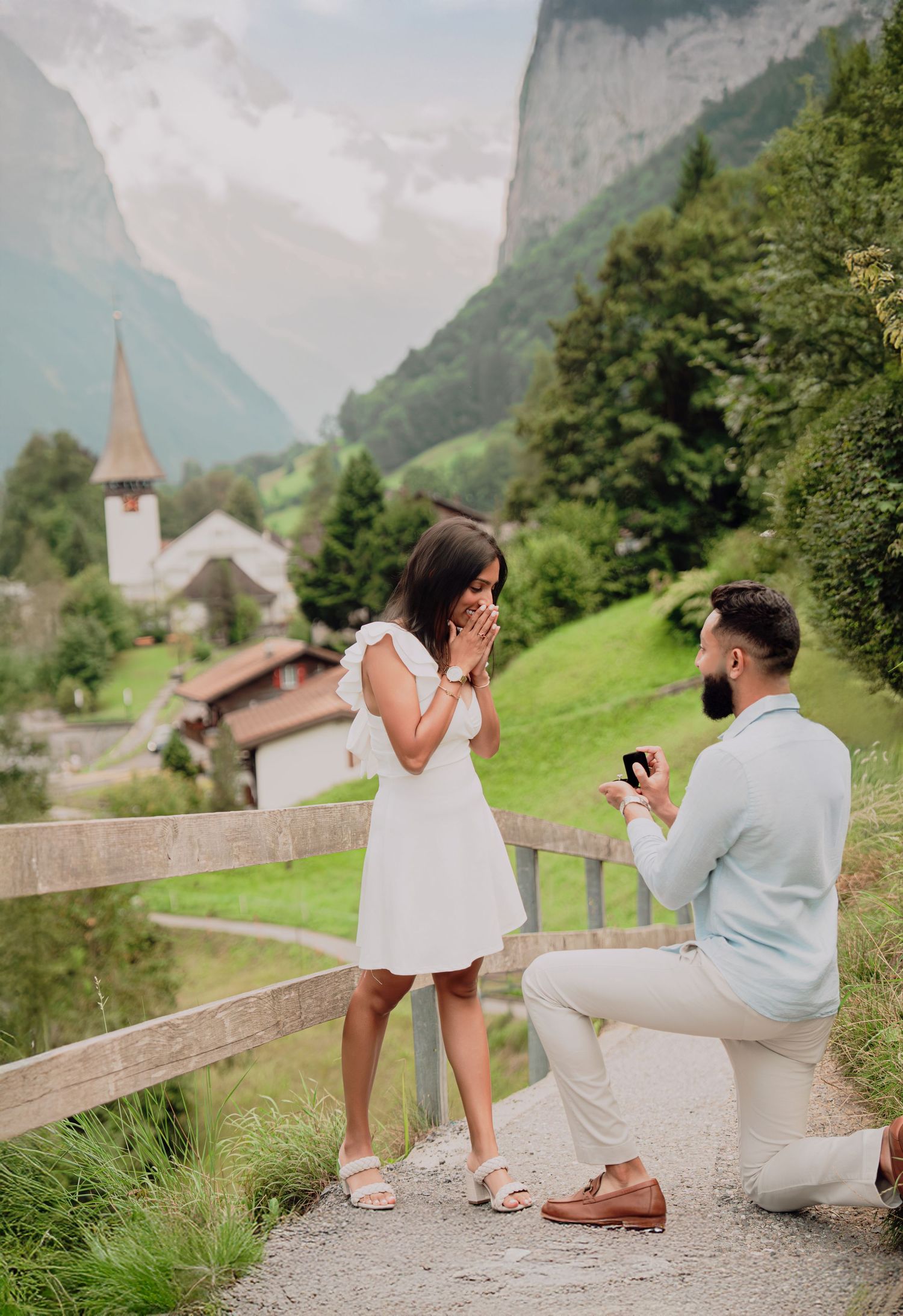 Woman covers mouth in surprise as man proposes on one knee in a Swiss alpine village.