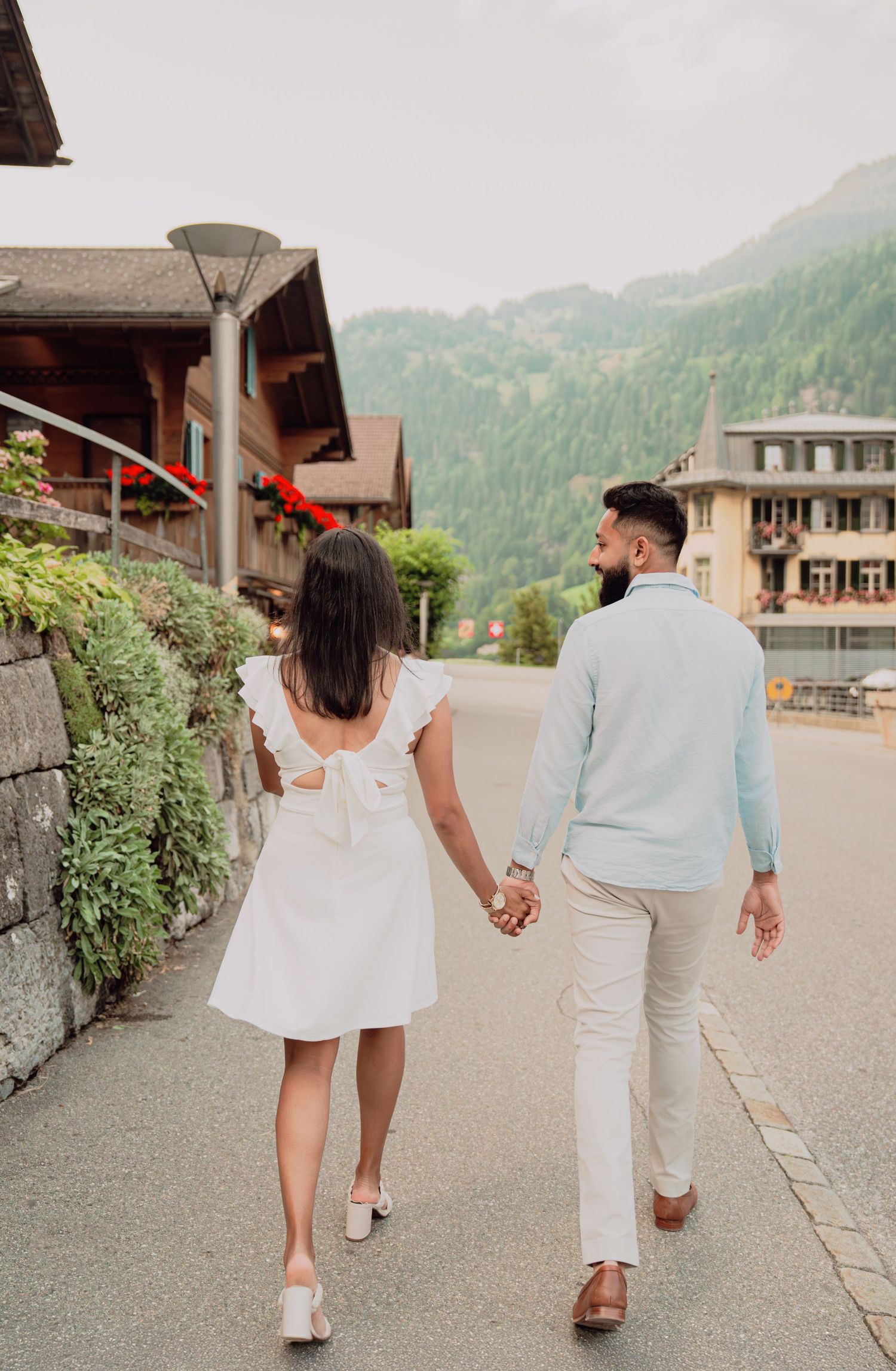 Couple walking hand in hand through a picturesque Swiss alpine village street surrounded by lush green mountains.
