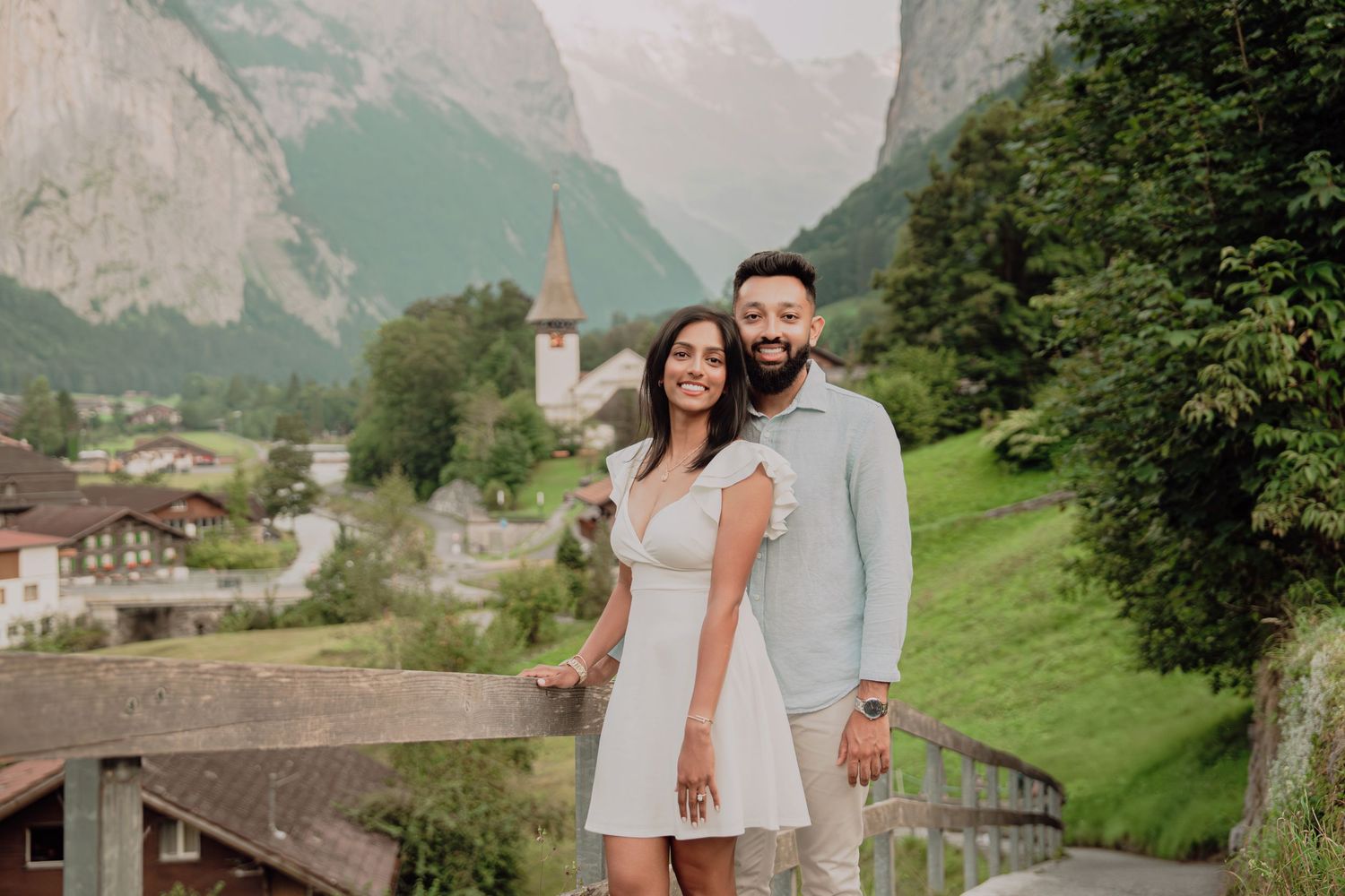 Happy couple embracing on bridge overlooking picturesque Swiss alpine village with church and mountain valley scenery.