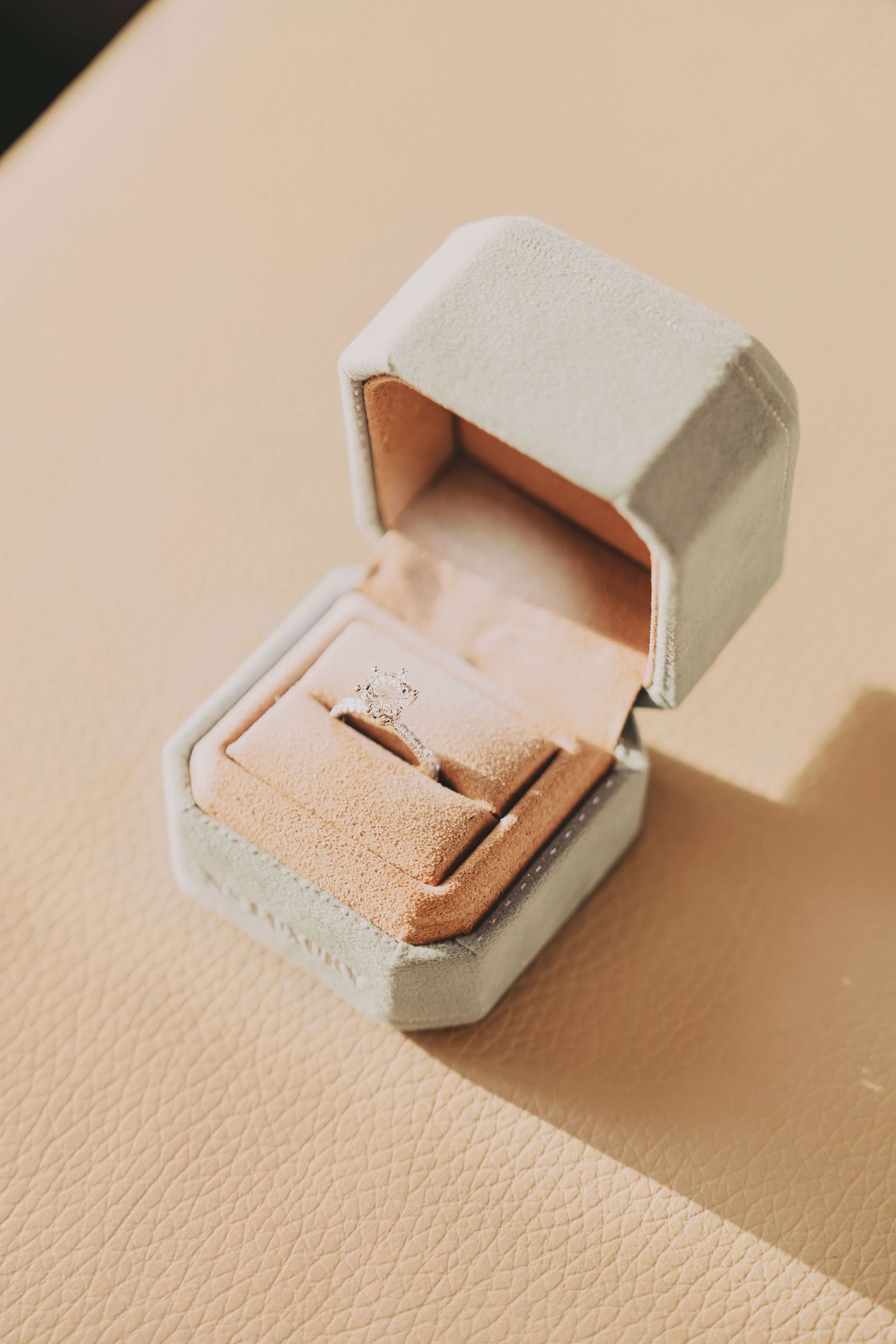 Elegant diamond solitaire engagement ring displayed in an open light gray velvet ring box on a beige surface.