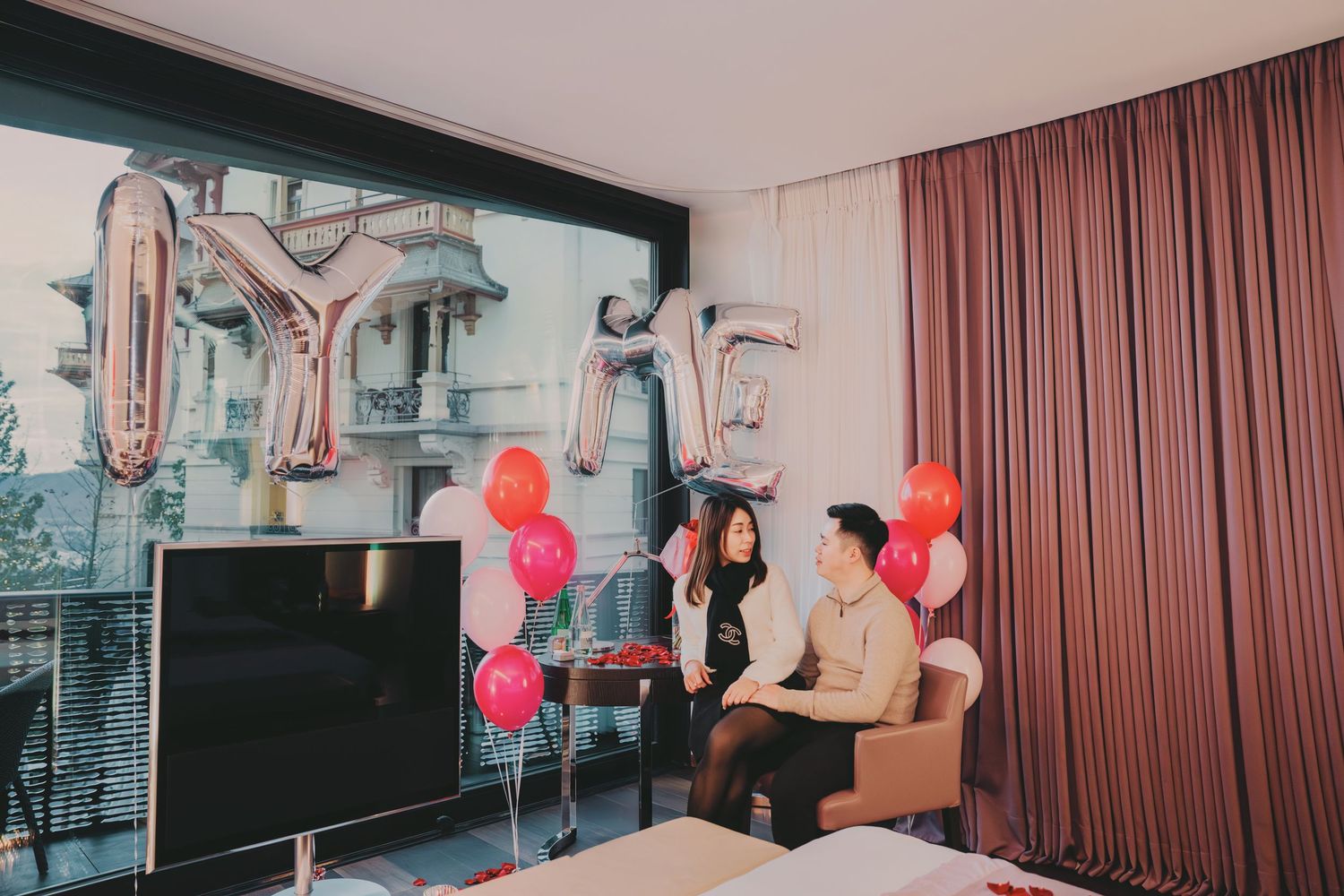 Couple seated in hotel room with LOVE silver letter balloons and colorful balloons by large window.