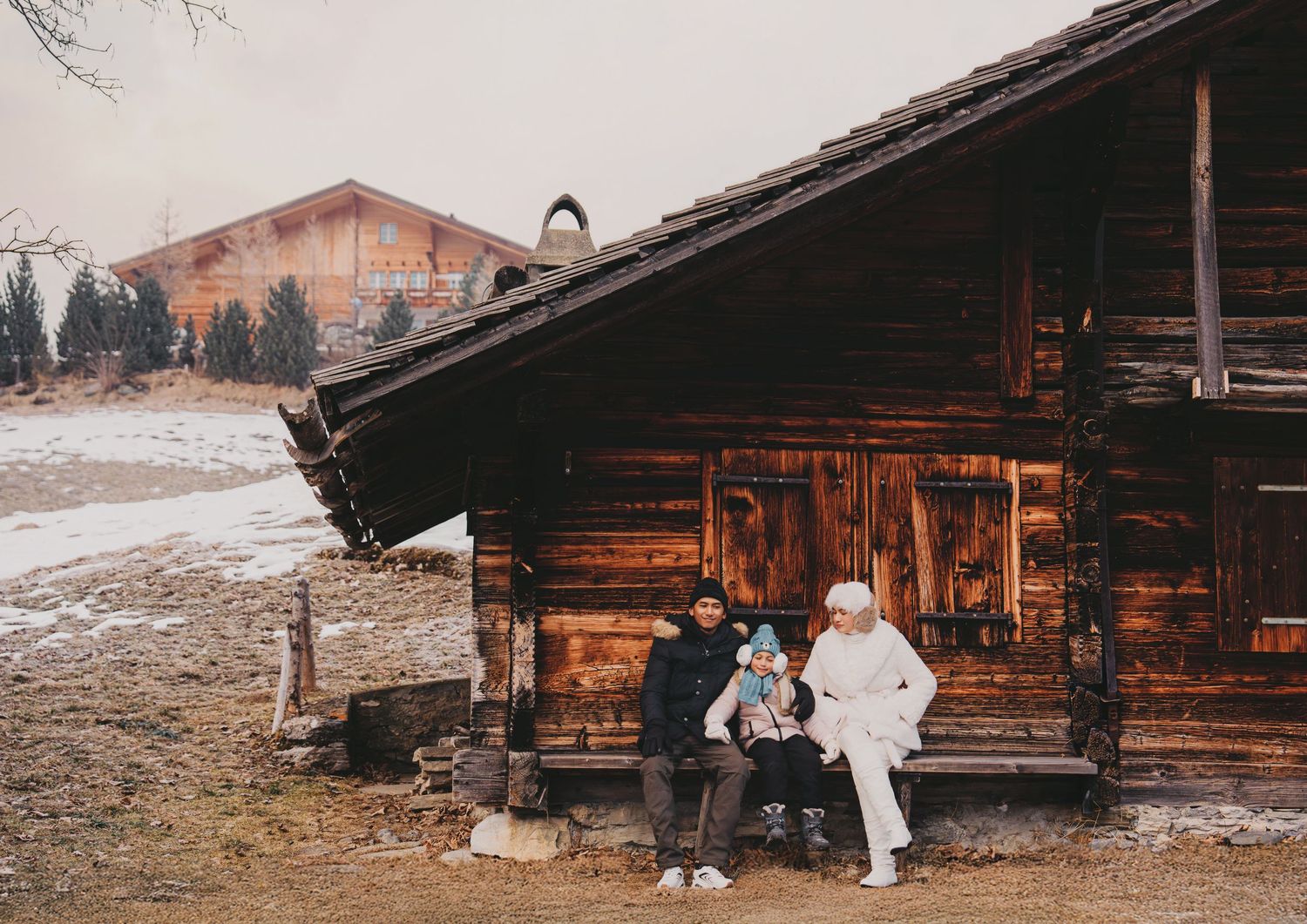 Family of three sitting on bench outside rustic wooden alpine cabin in snowy winter setting.