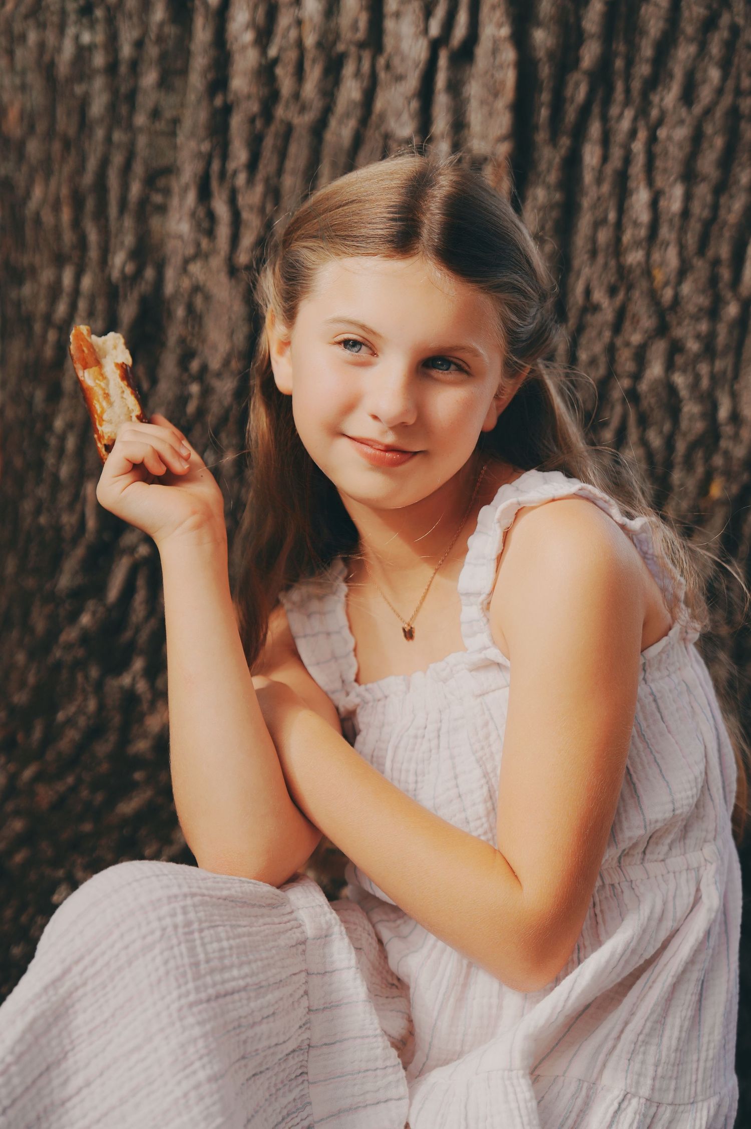 Young girl in white dress smiling against tree bark holding a snack outdoors.