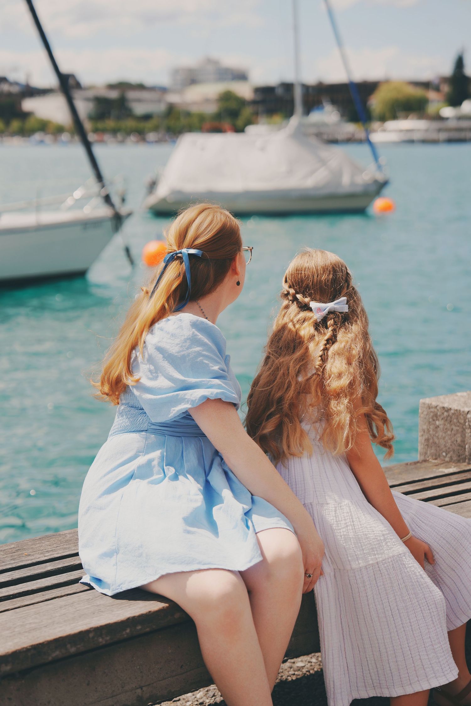 Two girls in summer dresses sitting on pier looking at sailboats on turquoise lake.