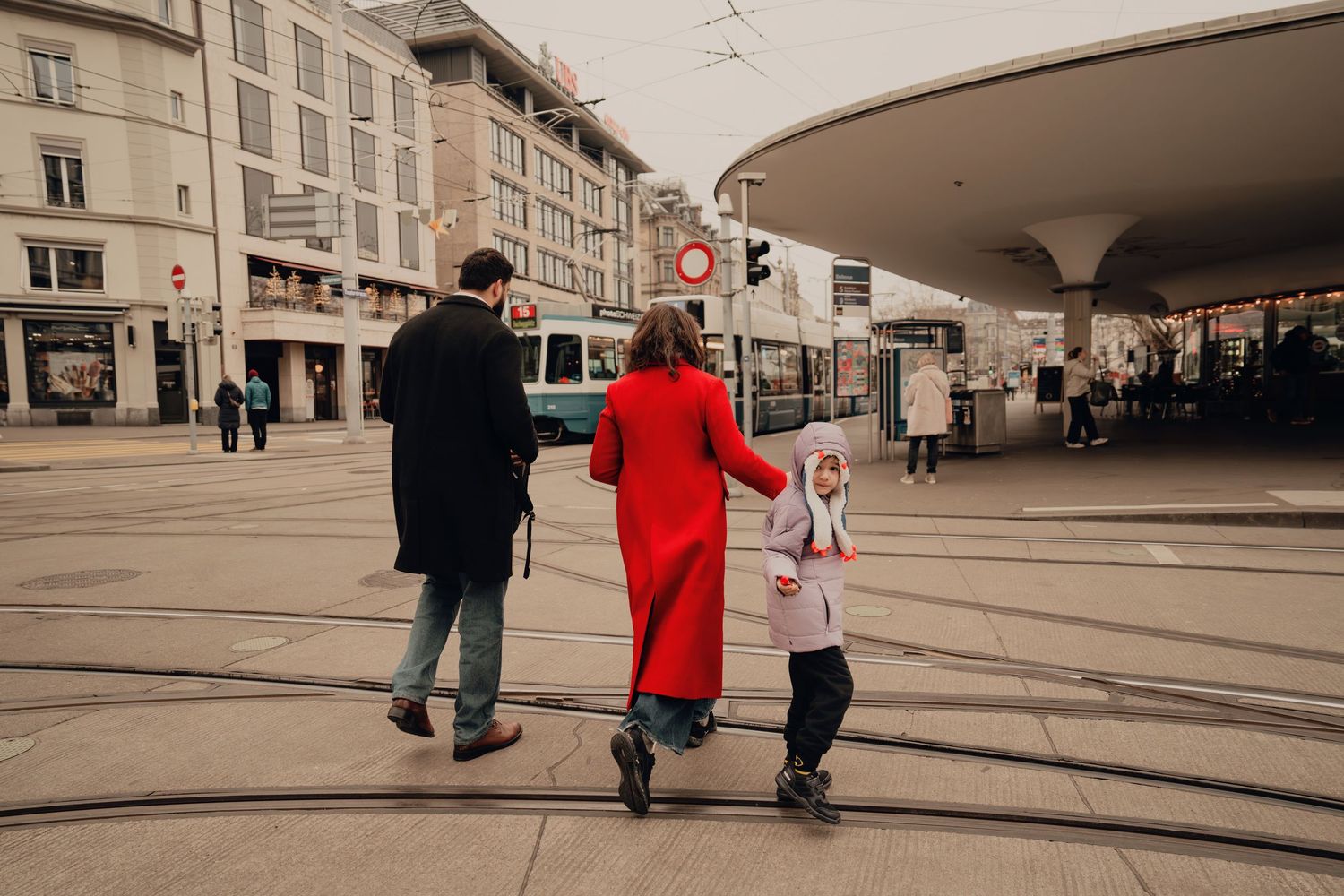 Family of three seen from behind crossing tram square in European city, woman in red coat.