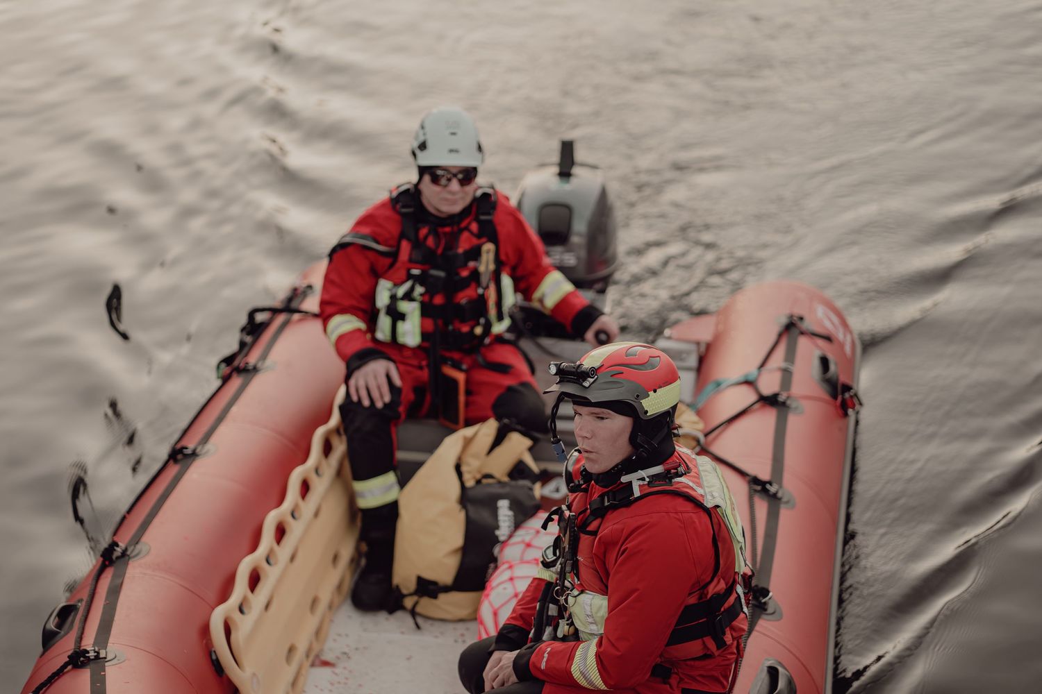 Two rescue workers in red suits operating an inflatable rescue boat on open water.