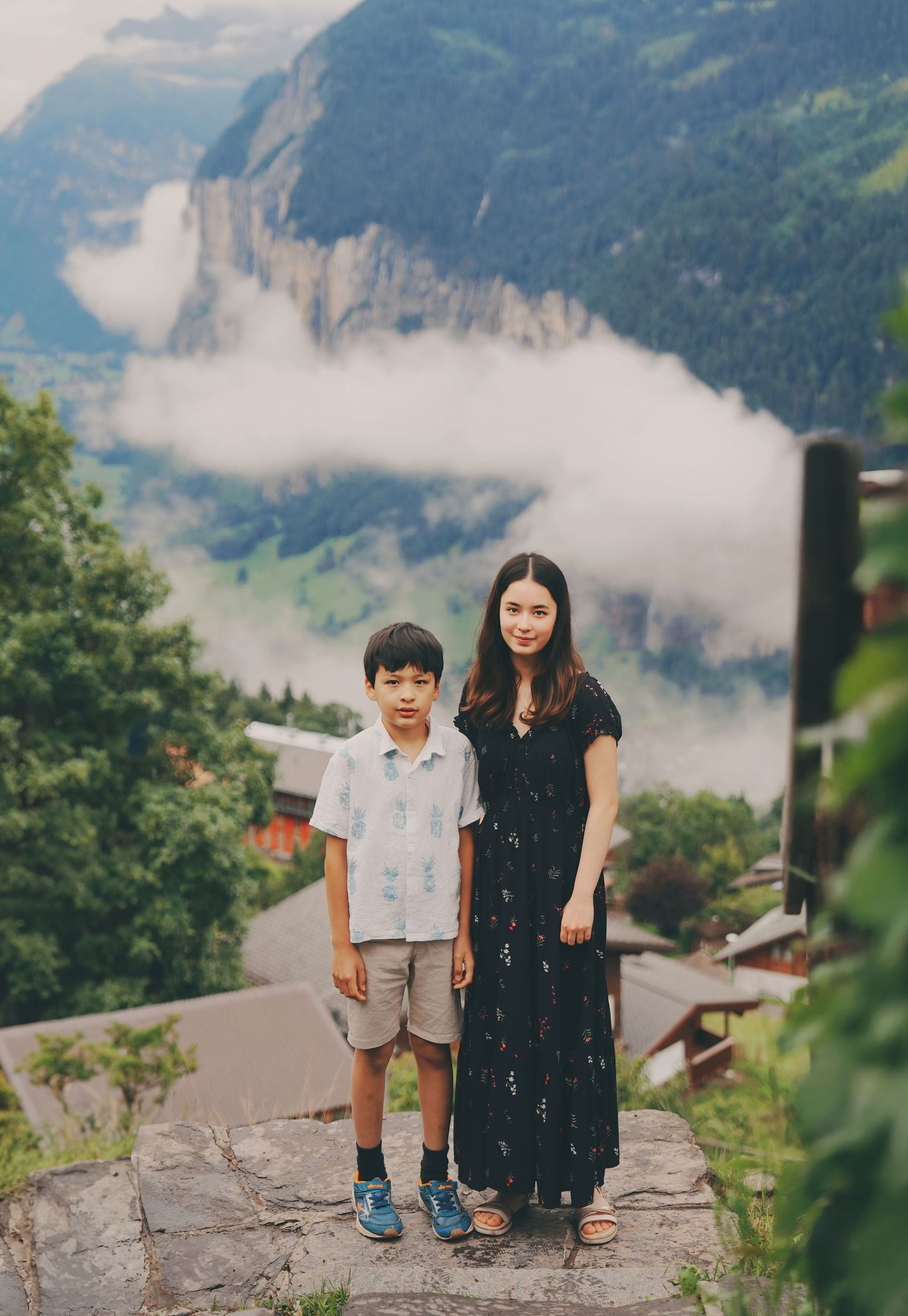 Mother in black dress and young boy in white shirt posing together on stone path with Swiss Alps in background.