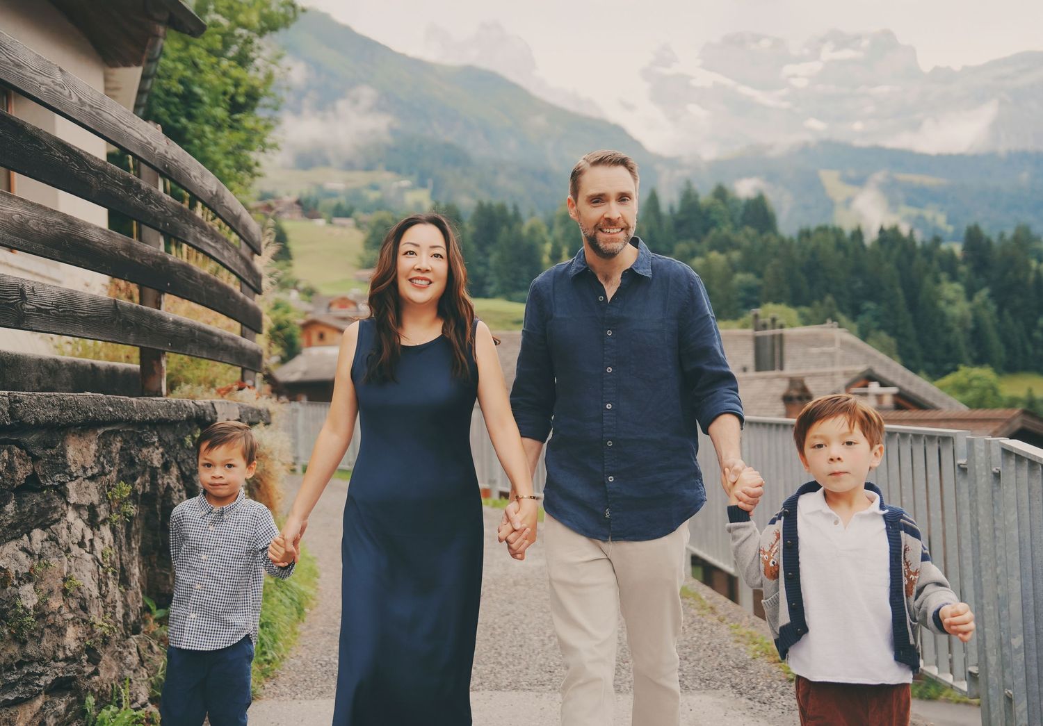 Happy family of four walking hand in hand on a scenic Alpine village path with mountains behind.