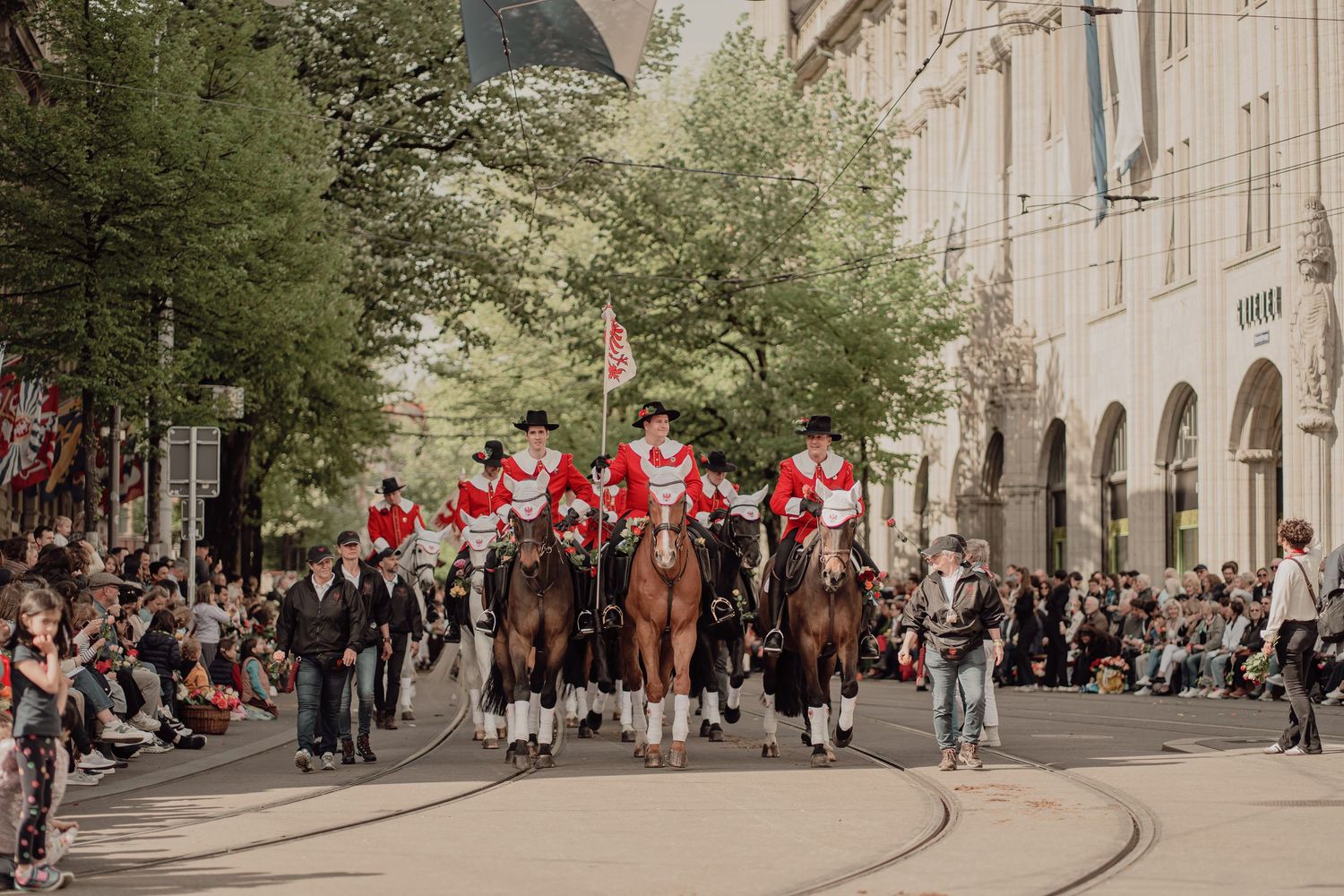 Riders in red and white Swiss costumes on horseback parading through tree-lined street.