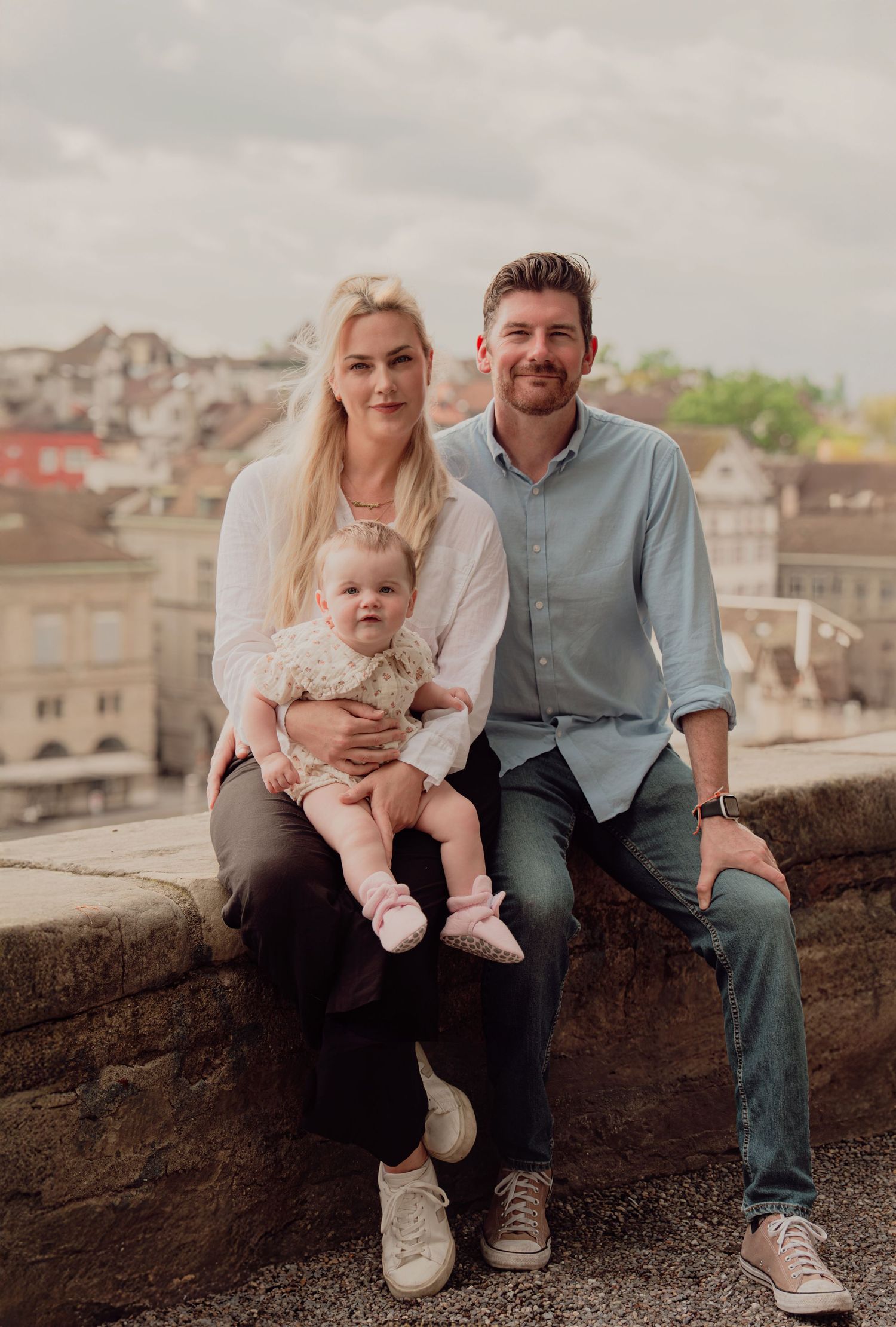 Young family with blonde mother, father, and baby sitting on stone wall in Zurich.