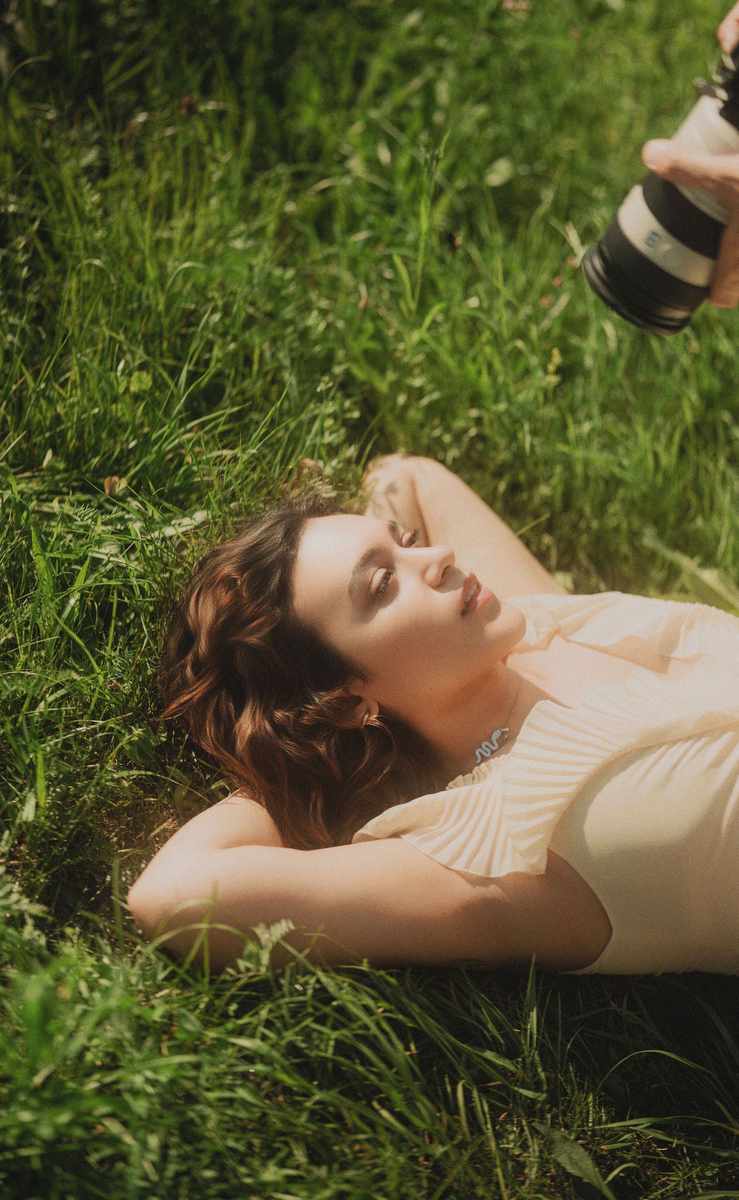 Woman in cream dress lying in green grass with eyes closed while photographer captures her portrait.
