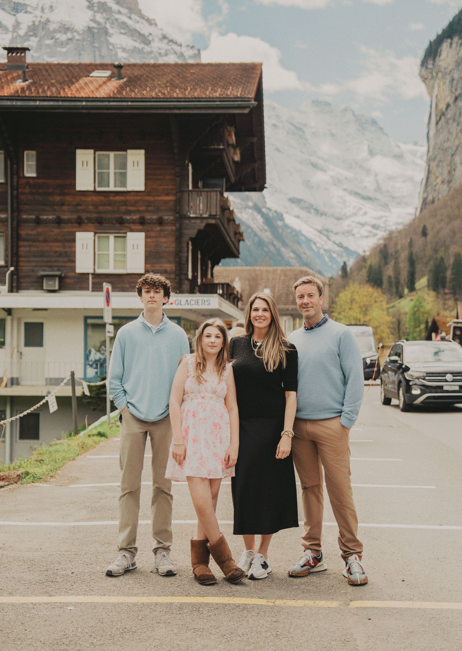 A family of four stands together on a village street with traditional Swiss chalets and snowy mountains backdrop.