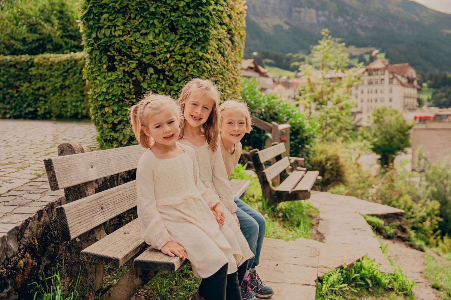 Three little girls in white dresses sitting on a bench in a Swiss alpine village.