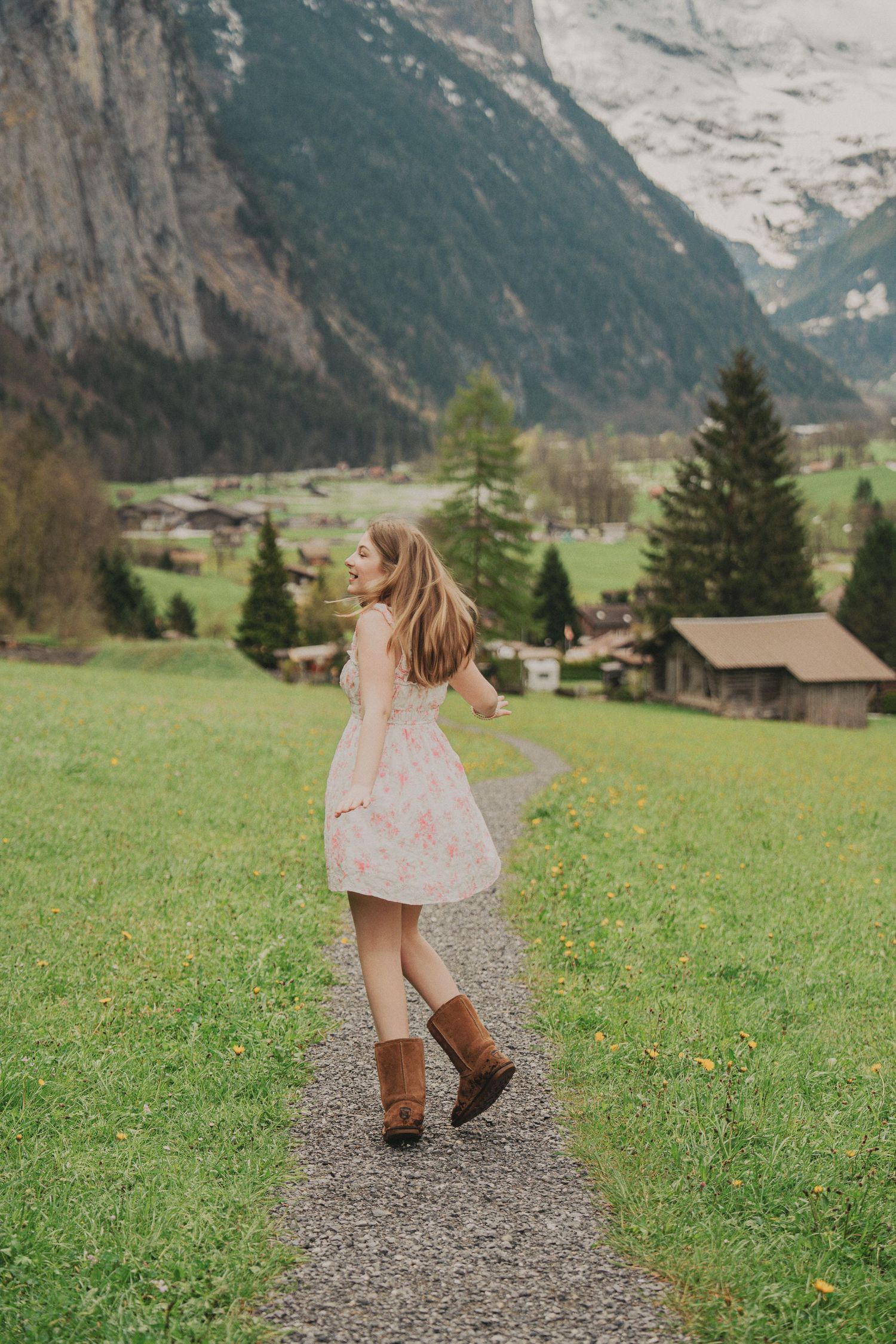 Young woman in floral dress and brown boots twirling on mountain path in Swiss alpine valley.