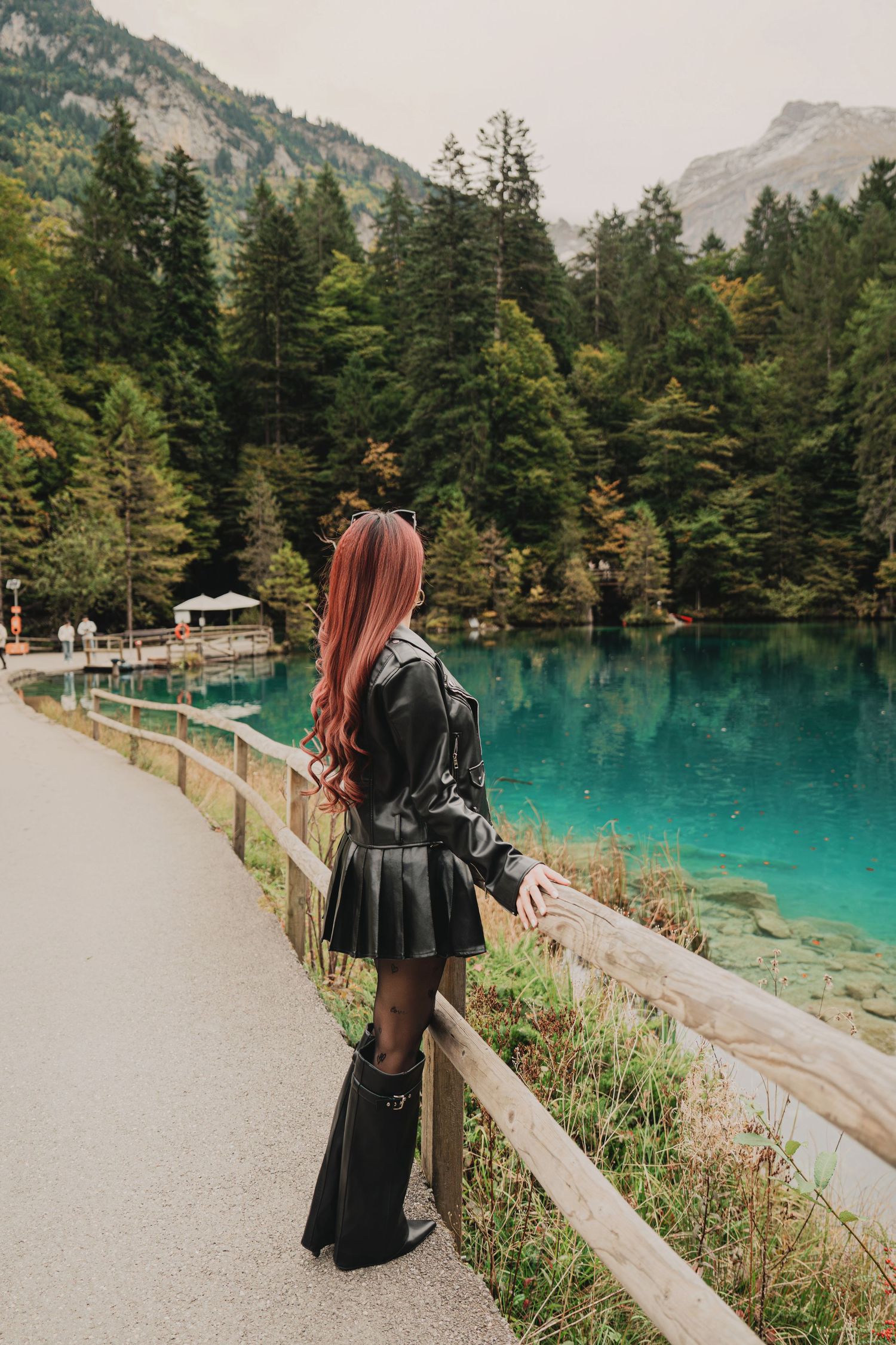 Woman with long red hair in black leather outfit gazing at stunning turquoise alpine lake and forest.