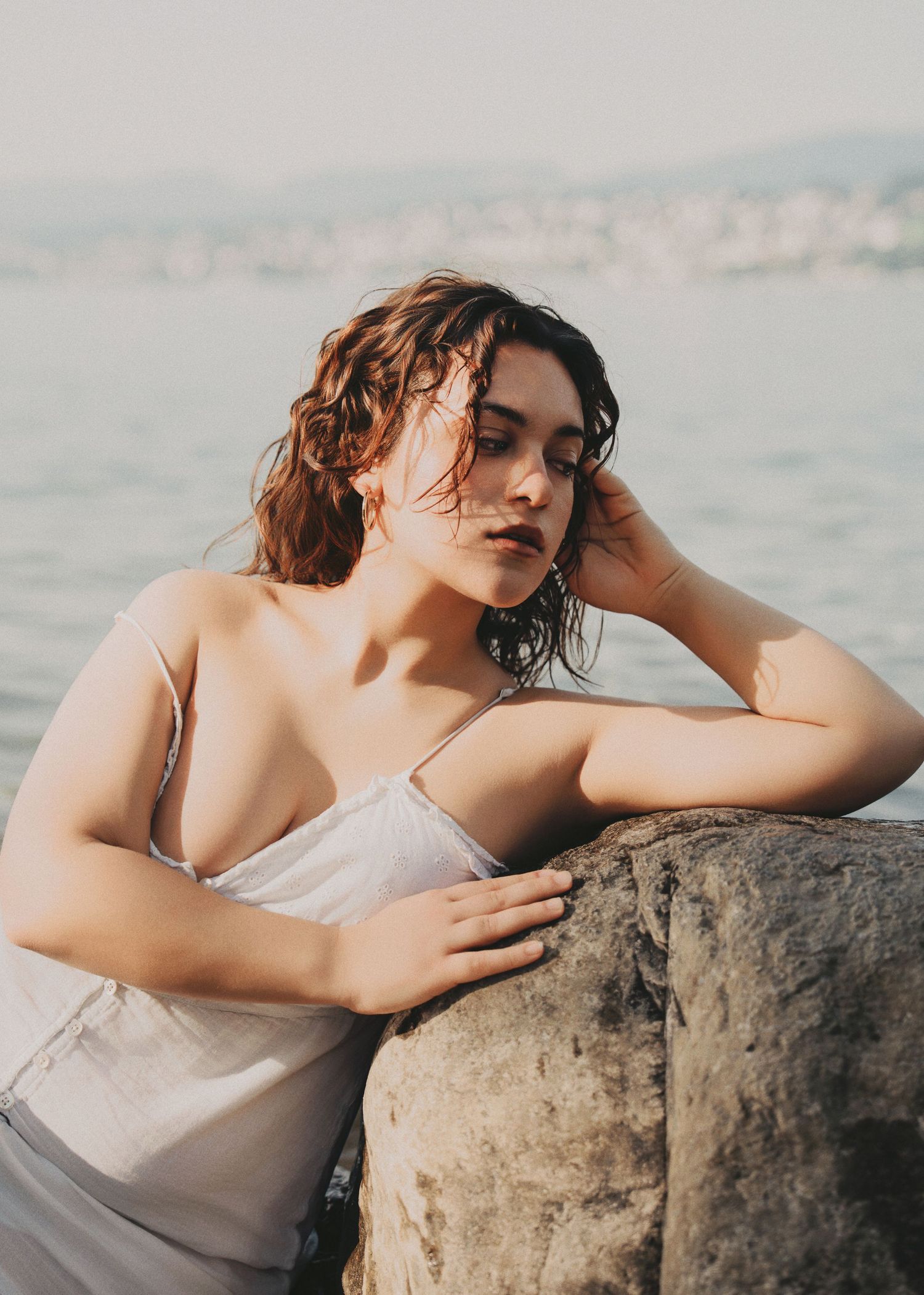 Brunette woman in white dress leaning on coastal rock with sea behind her.
