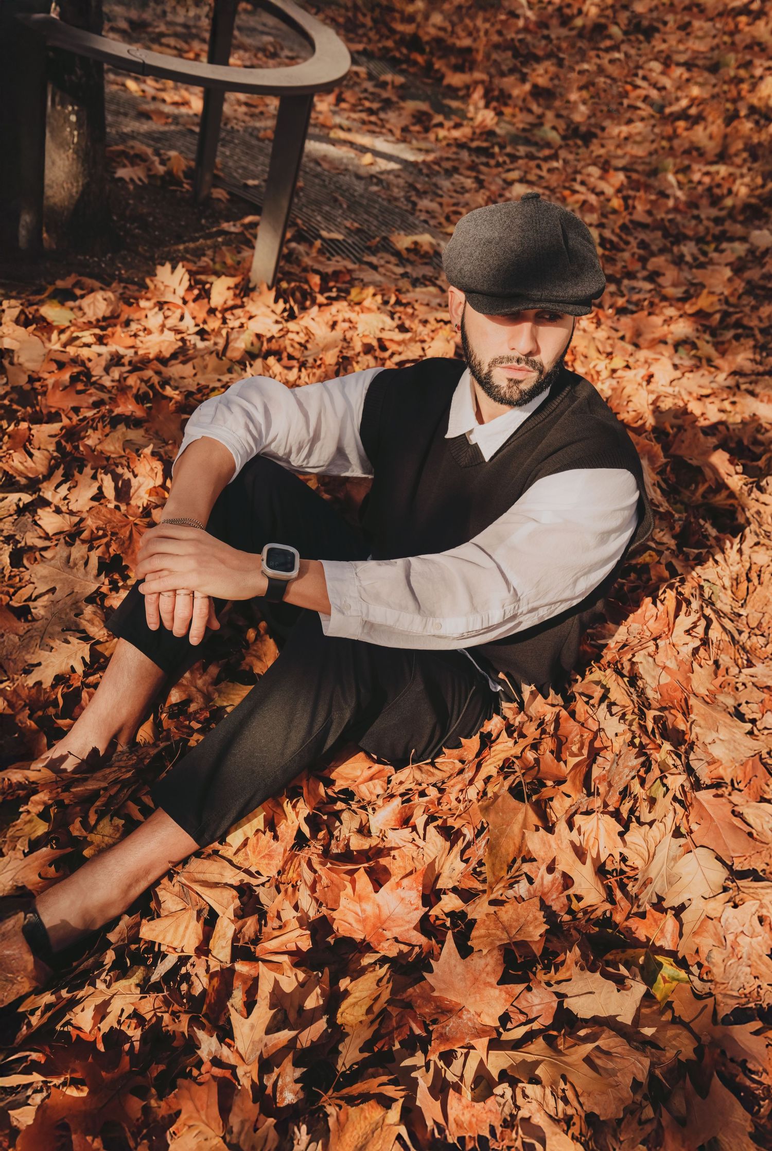 Bearded man in vest and flat cap relaxing in golden autumn leaves with smartwatch visible.