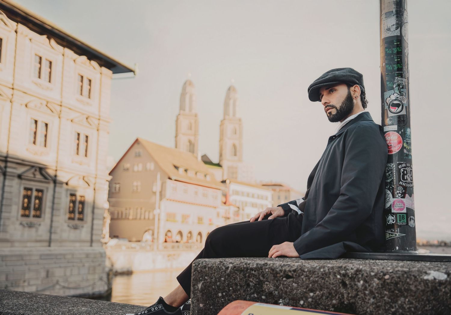 Bearded man in black coat and flat cap sitting on stone wall with Zurich cityscape in background.