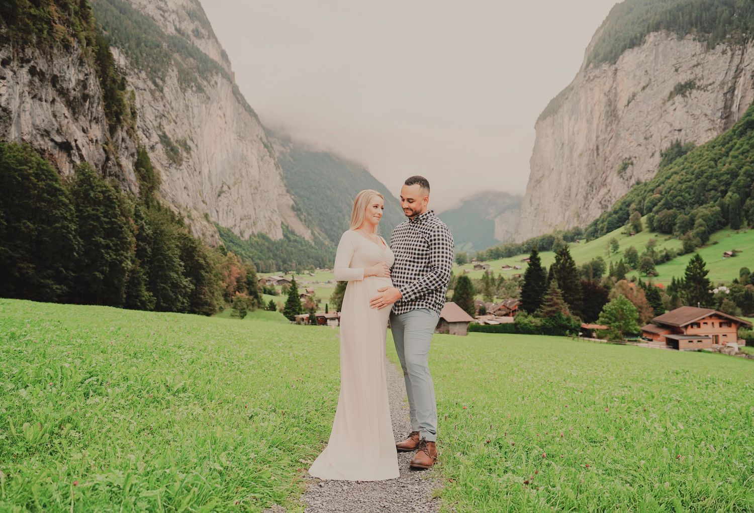 Pregnant couple posing together on a mountain valley path in Switzerland, woman in white gown, man in plaid shirt.