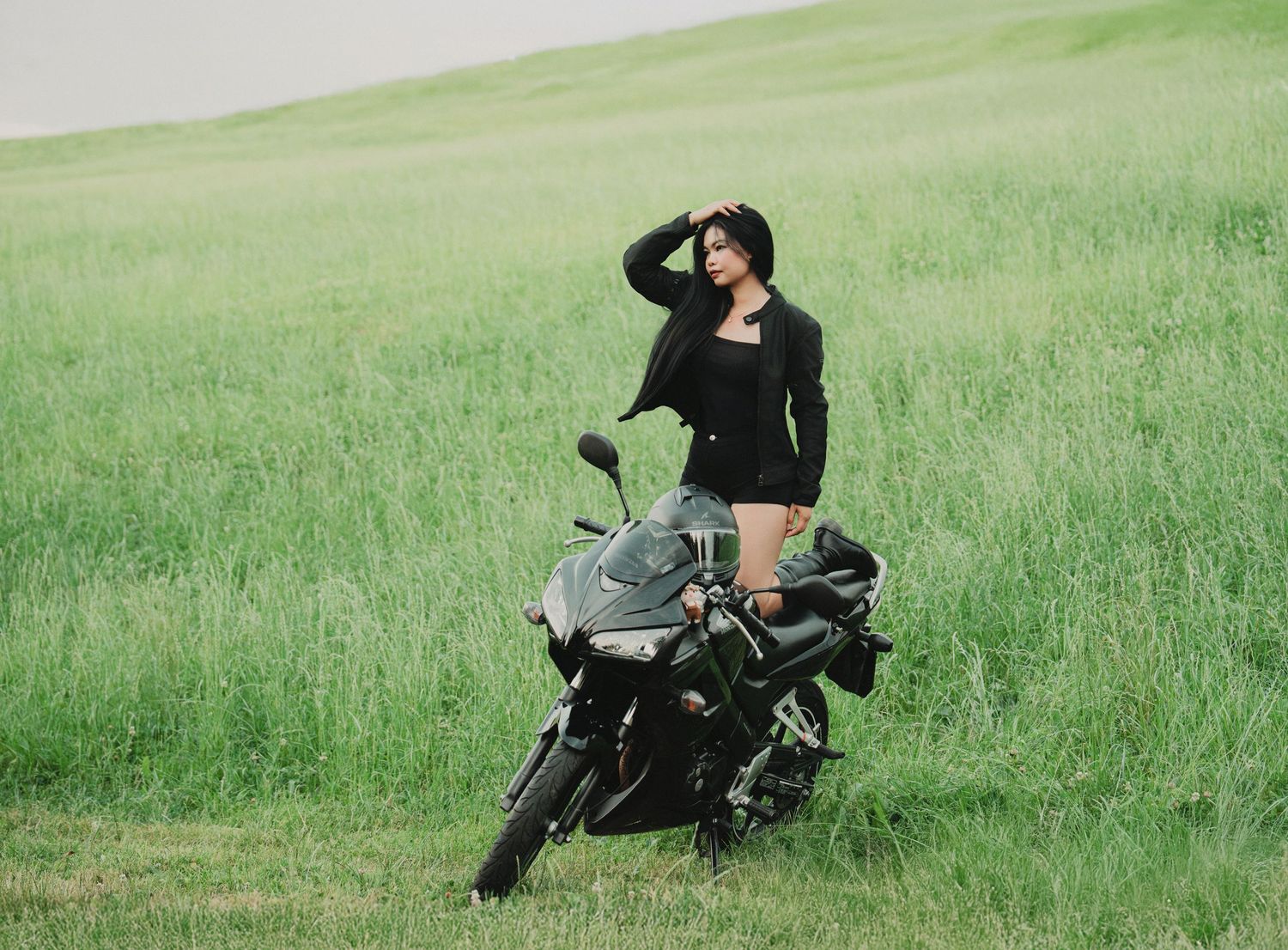 Woman in black outfit leaning on a black sport motorcycle on a scenic green meadow.
