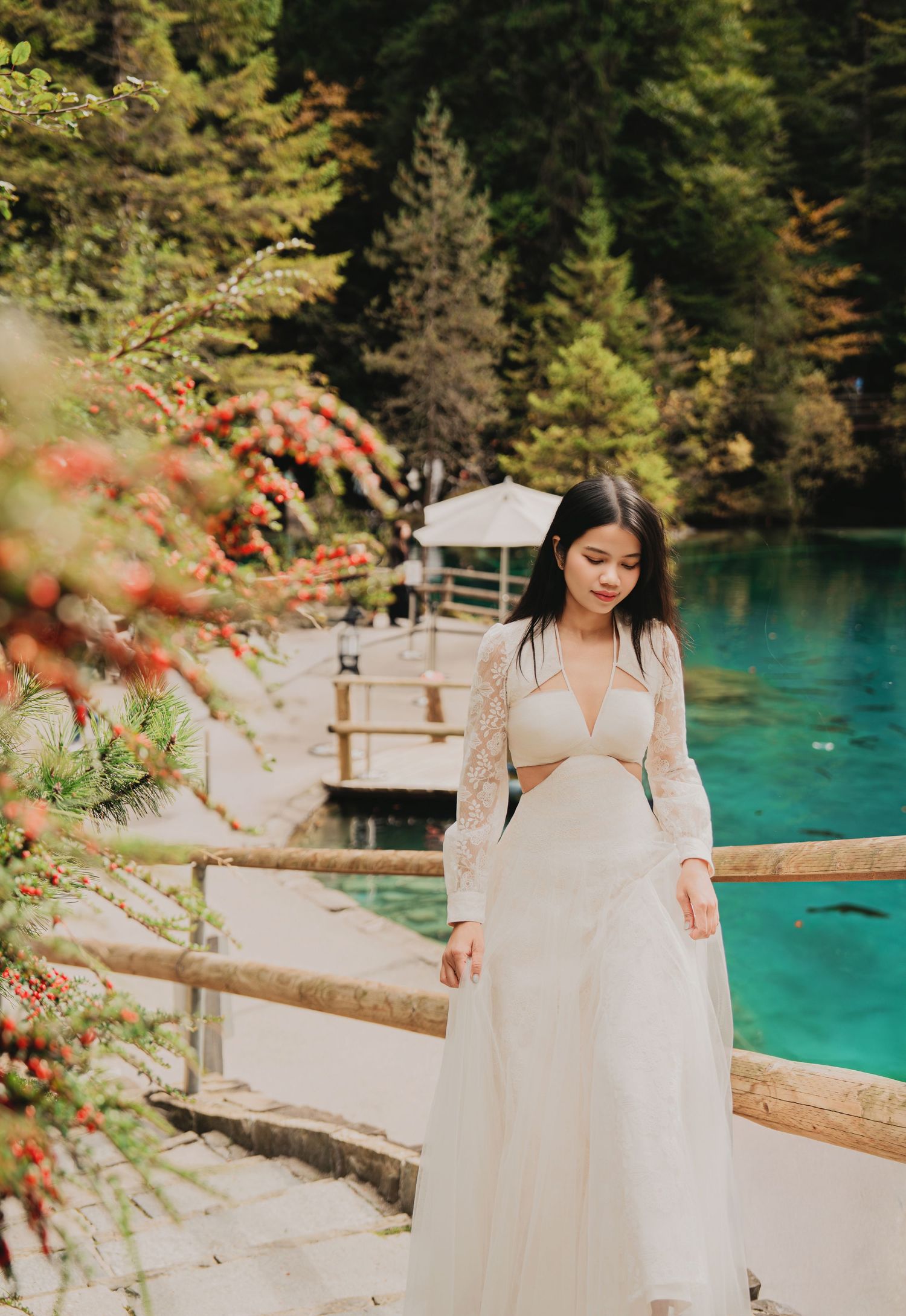 Woman in white lace two-piece dress walks near turquoise lake with red berry bushes.
