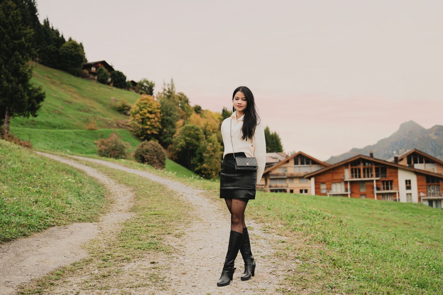 Young woman in cream sweater and black skirt walks along rural path with Alpine chalets in background.