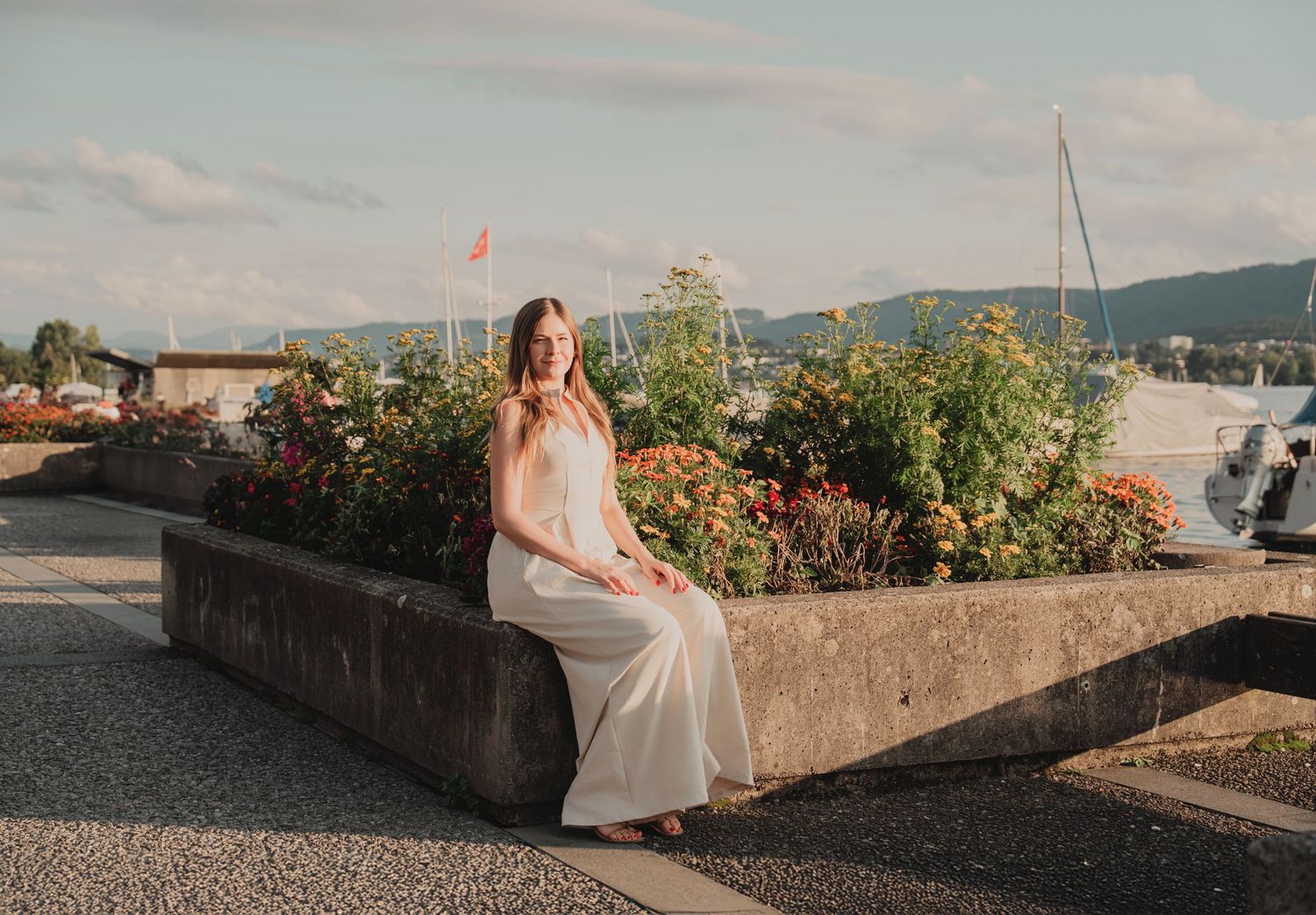 Woman in white dress sitting on stone planter with flowers and marina in background at golden hour.