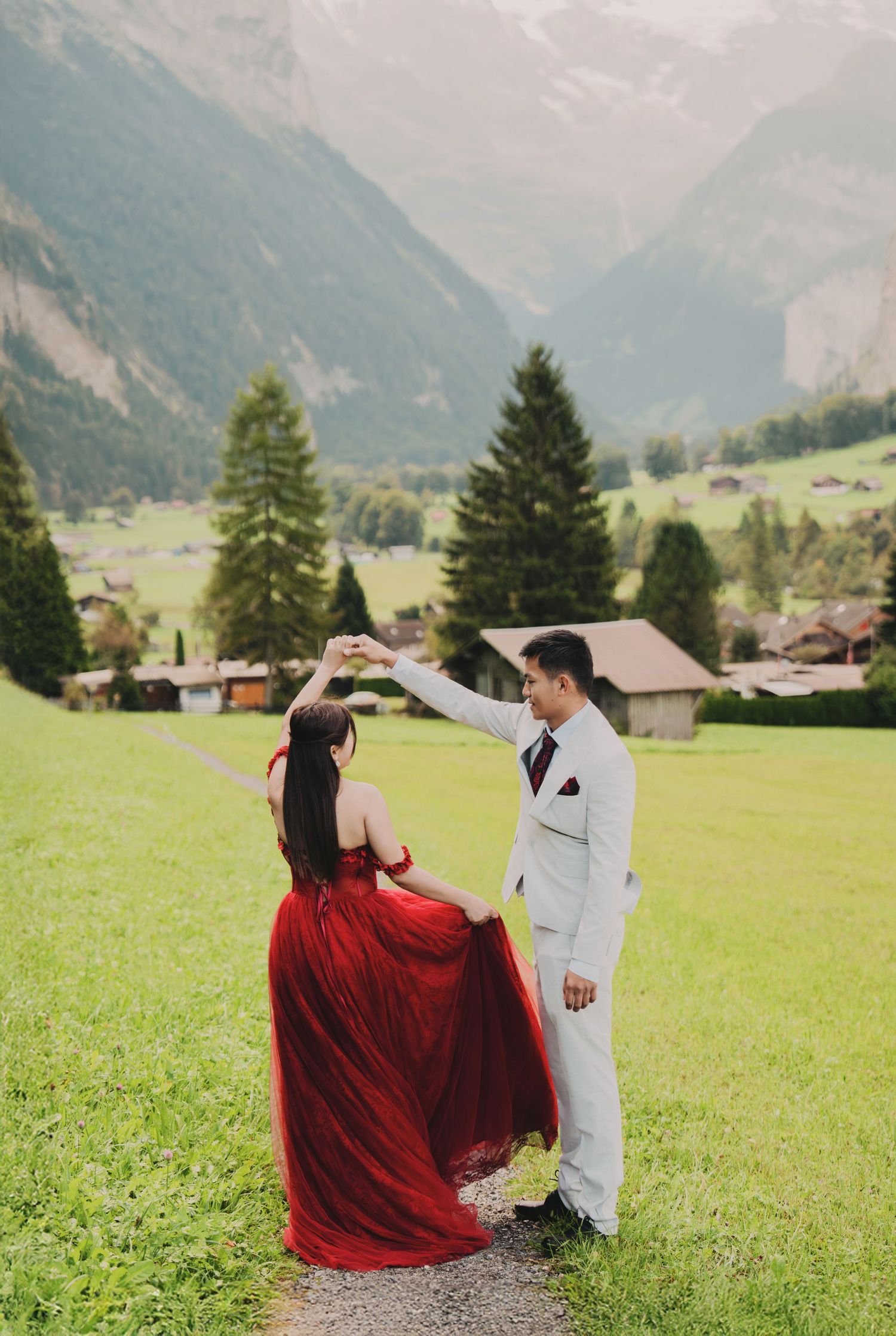 Groom twirling bride in red tulle gown on green meadow with Swiss mountain valley backdrop.