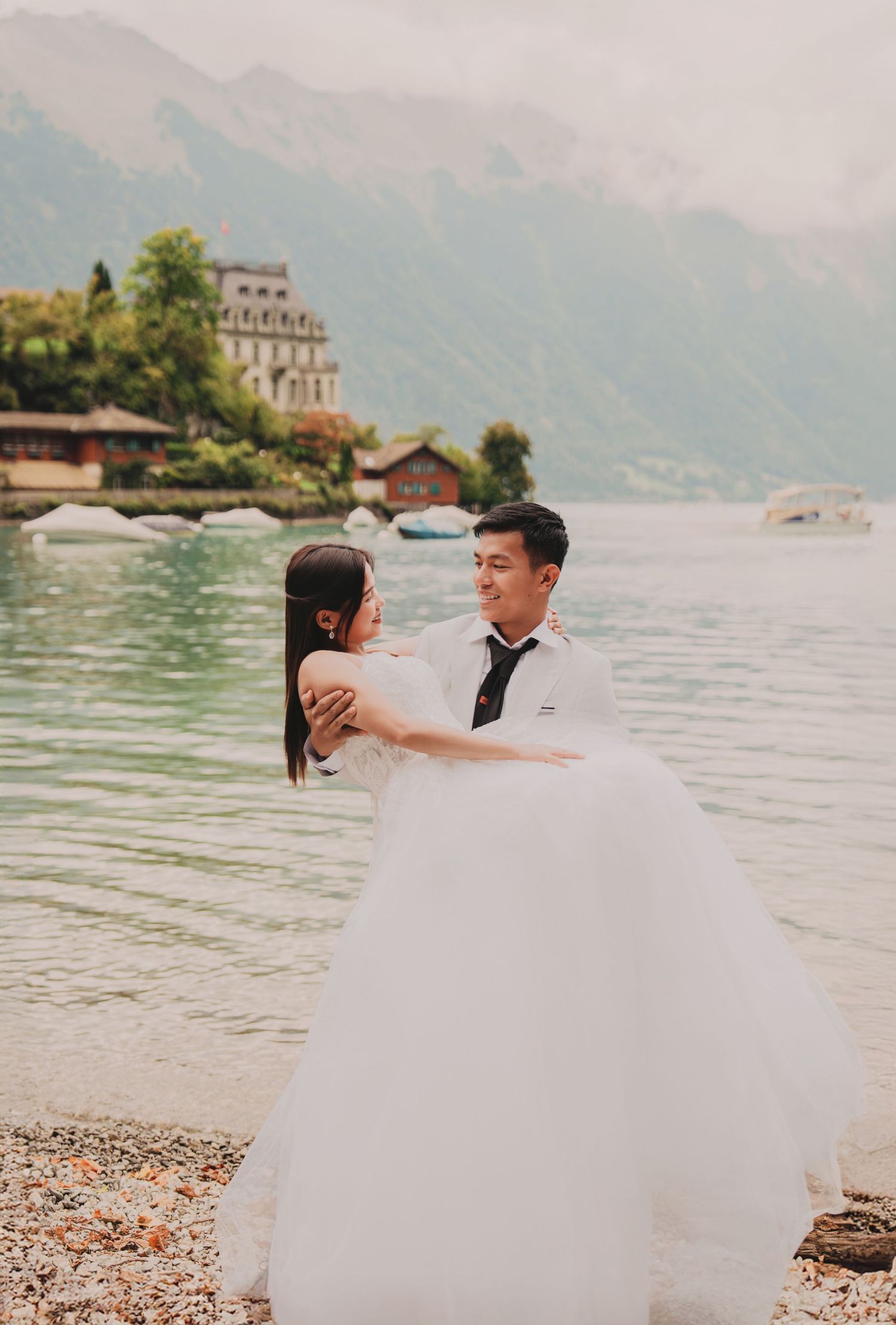 Couple in wedding attire embracing by a scenic alpine lake with mountains and historic buildings in background.
