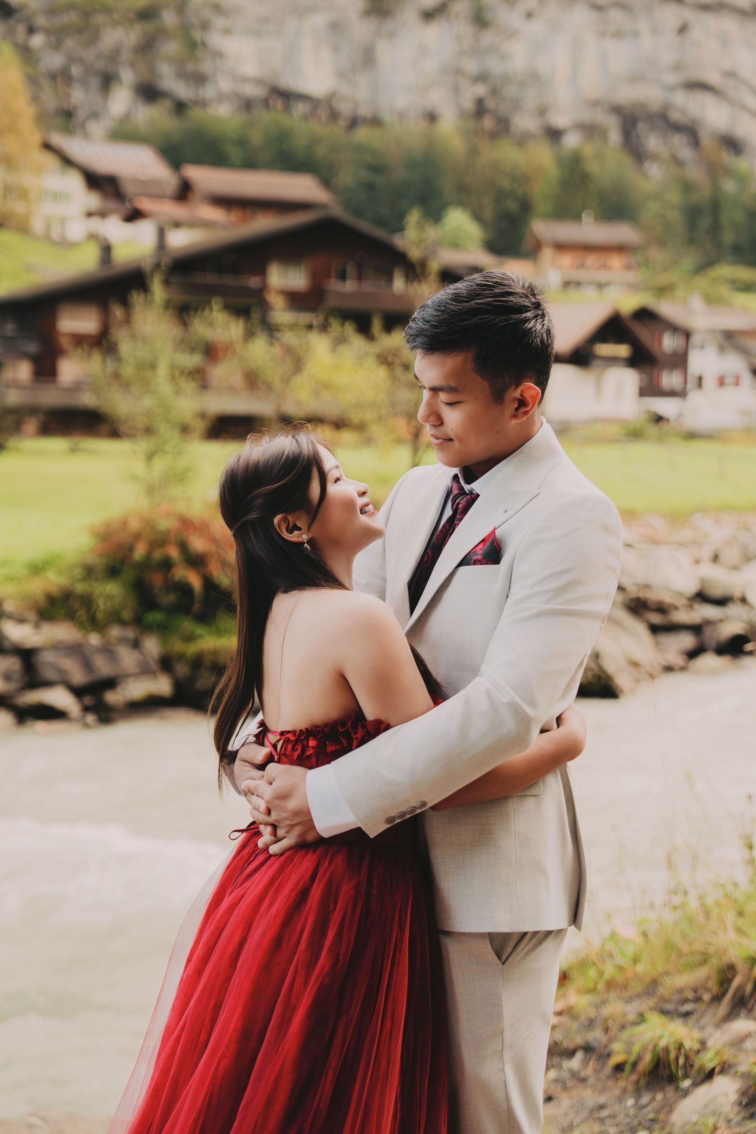 Laughing bride in red tulle gown embraced by groom in beige suit with Swiss village backdrop.