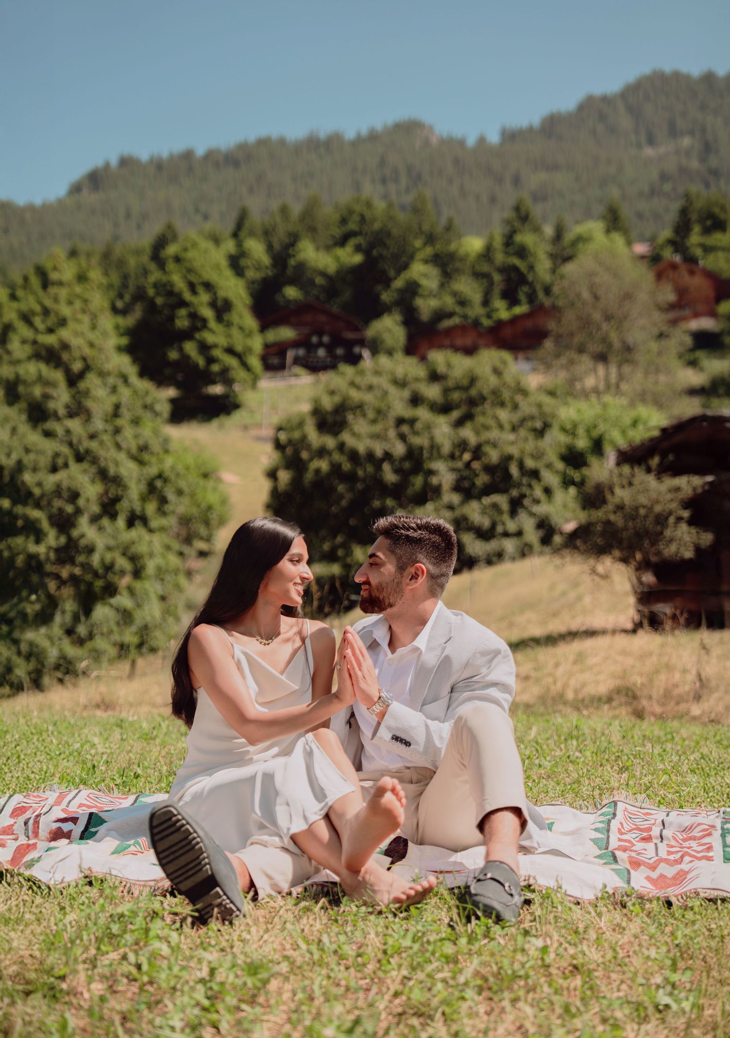 Happy couple sitting on a blanket in a sunny green meadow surrounded by lush forest and rustic buildings.